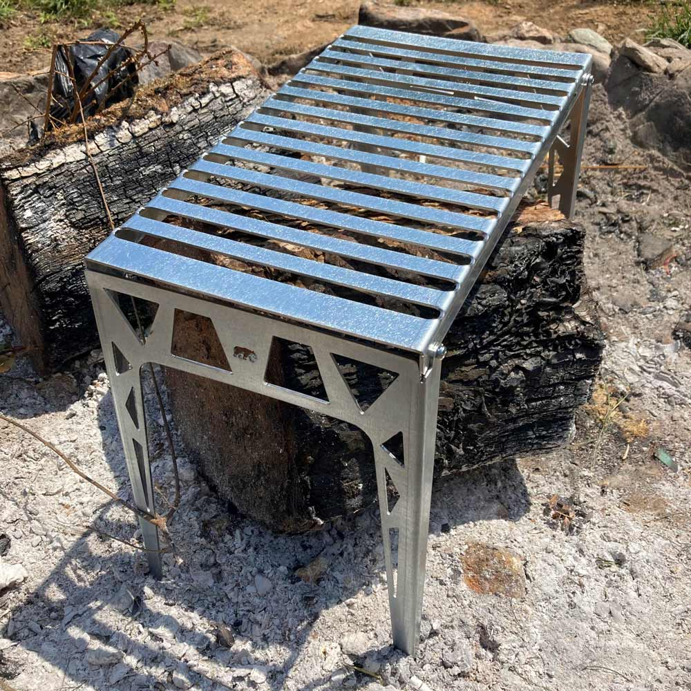 Metal bir ızgara dışarıda kömürleşmiş kütüklerin üzerinde duruyor. Izgaranın çıtalı bir yüzeyi ve üçgen destekleri var.