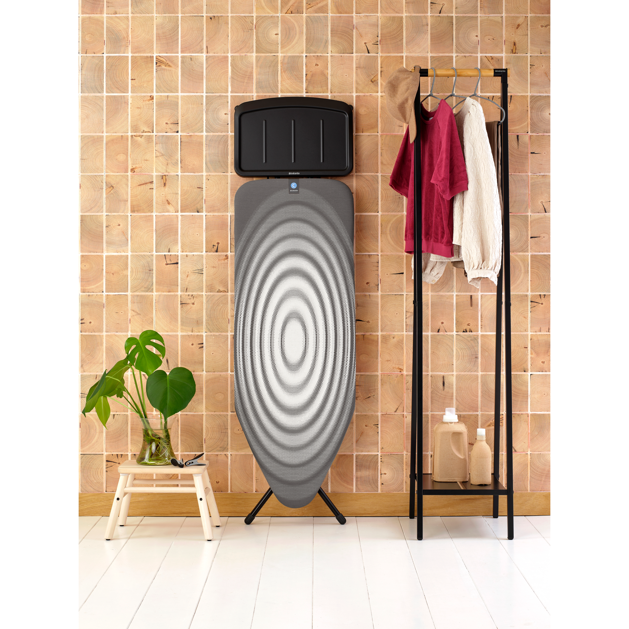 An ironing board with clothes and a plant on a stool.