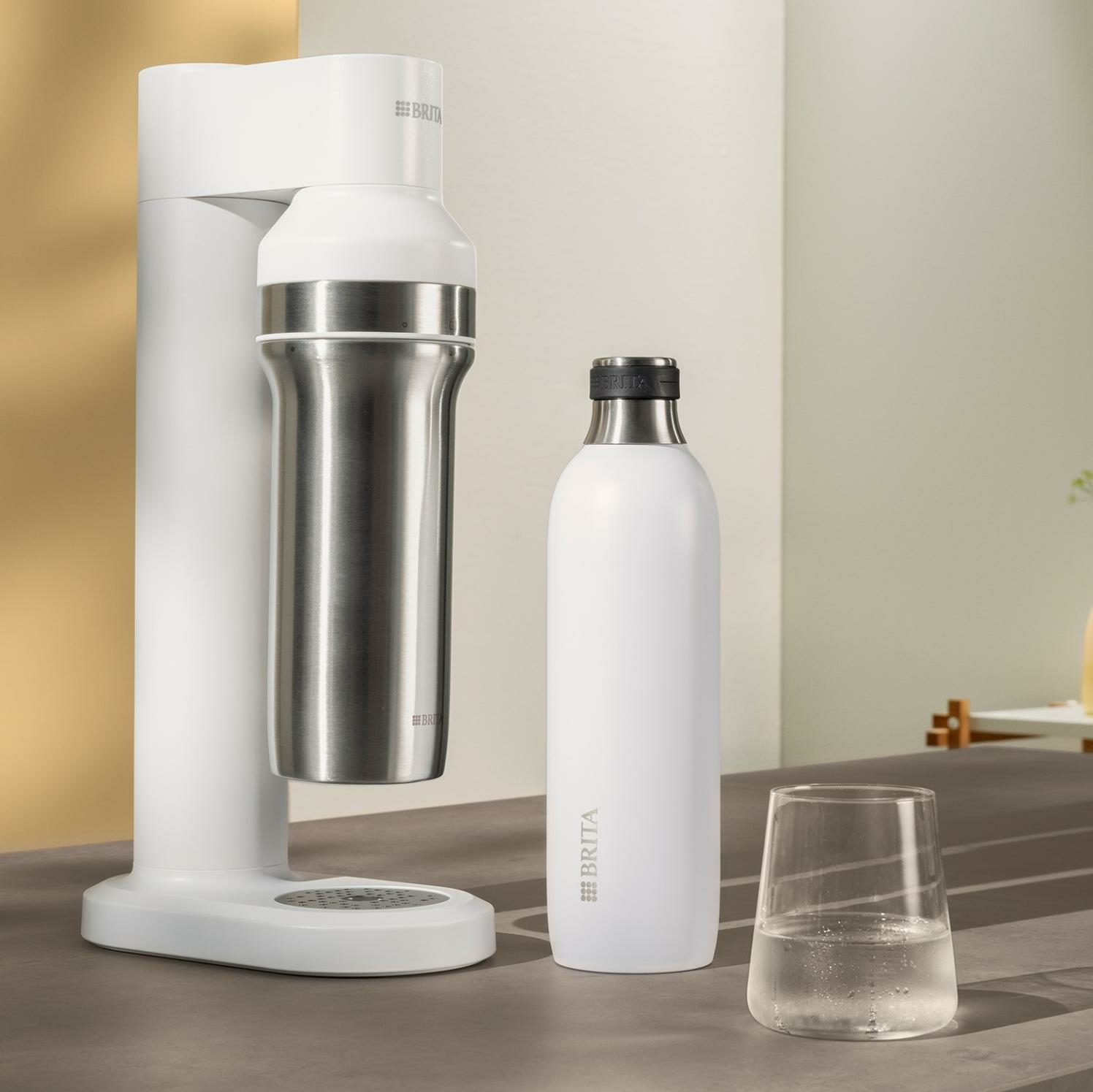 Ein weißer Brita Sodastreamer mit Edelstahlflasche, weißer Flasche und Wasserglas.