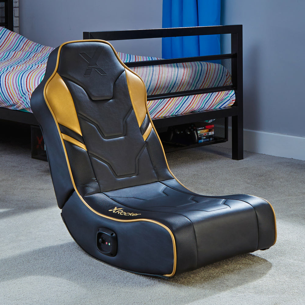 Ein schwarzer und goldener Gaming-Stuhl steht auf einem Teppich neben einem Bett.
