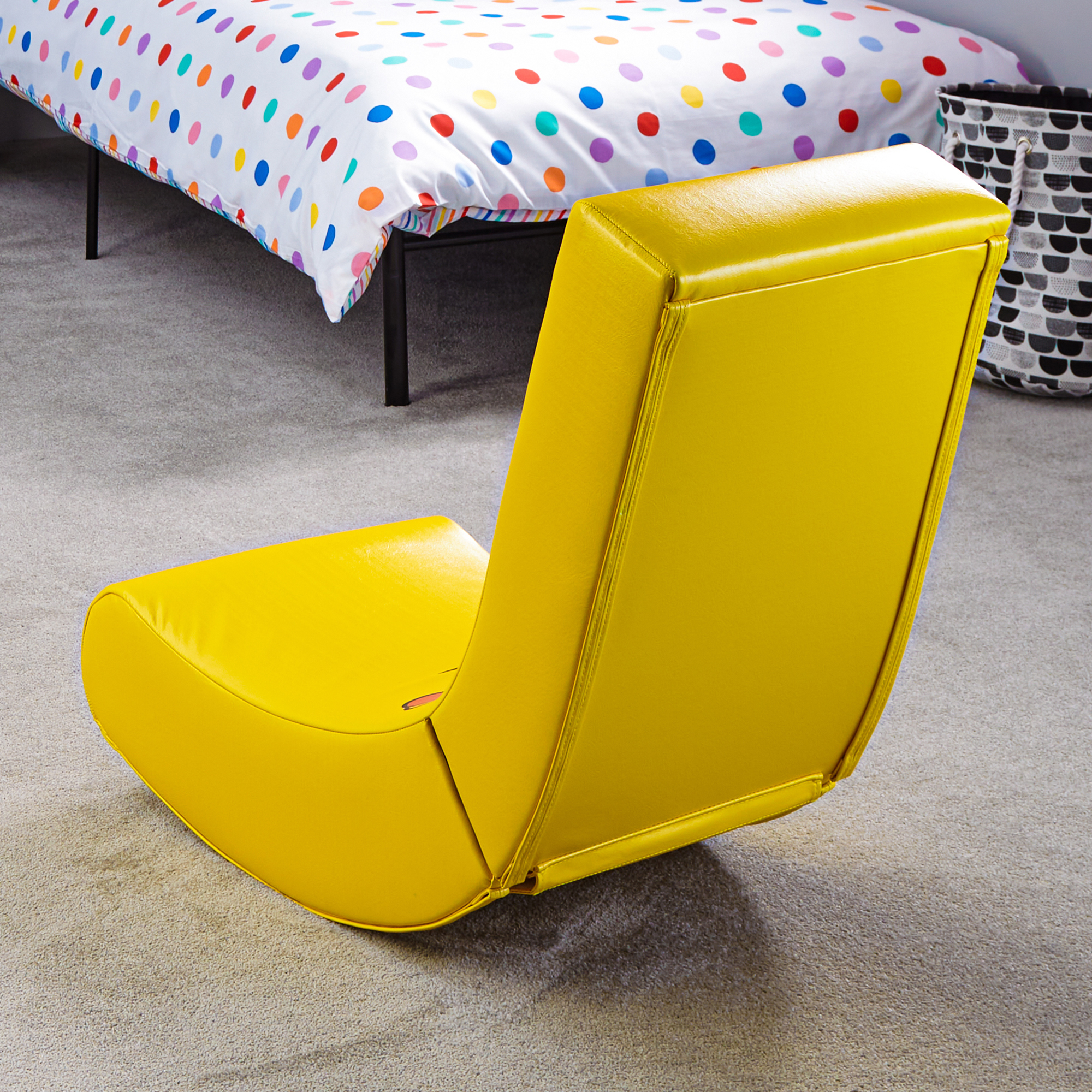 Een gele schommelstoel op een tapijt naast een bed met stippenpatroon.