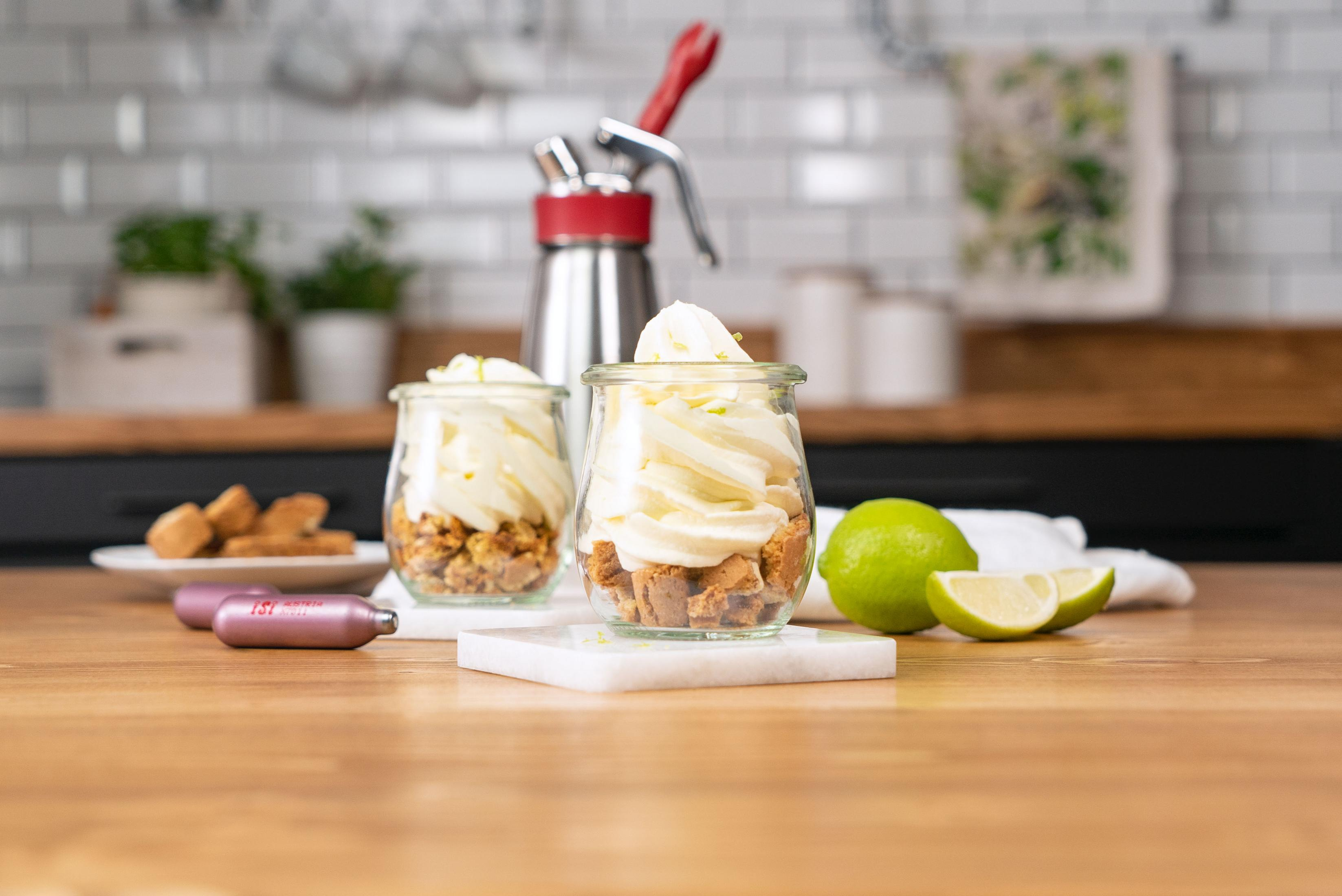 Zwei Gläser mit Sahne und Krümeln, Sahnebereiter, Limetten und Keksen auf einem Holztisch.