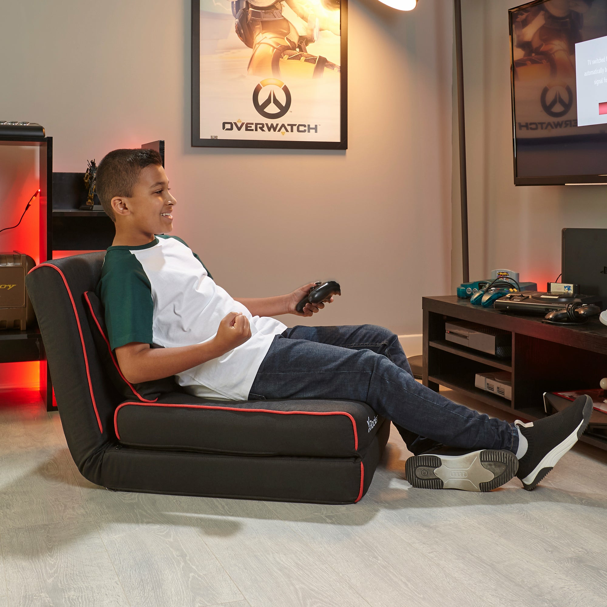 Ein Junge spielt mit einem Controller in einem Raum mit Overwatch-Poster.