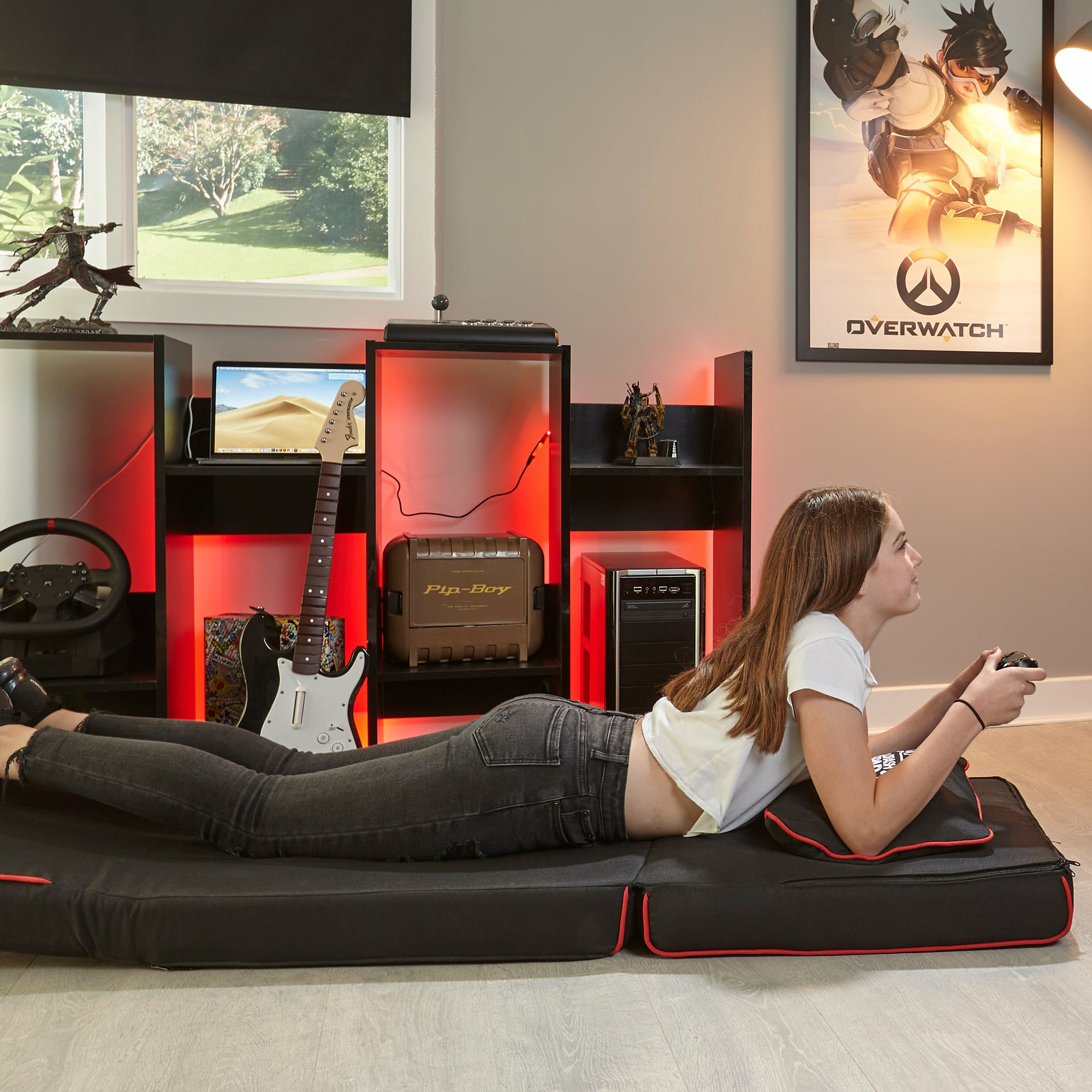 Mädchen liegt auf schwarzer Matte vor einem Gaming-Setup mit Gitarre, PC und Poster.