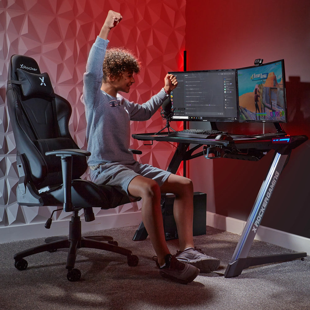 Ein junger Gamer hebt triumphierend die Arme in einem Gaming-Stuhl vor einem Schreibtisch mit Monitoren.