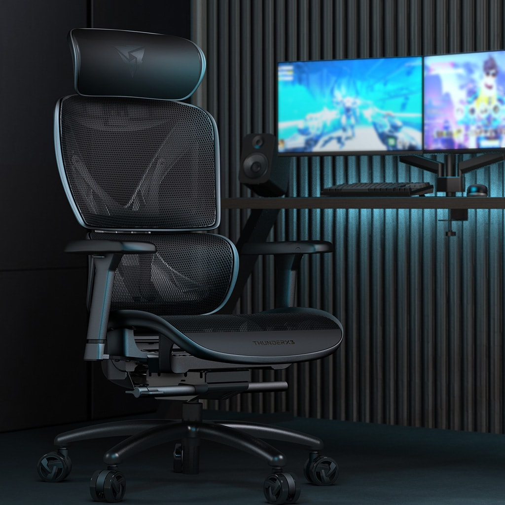 Silla gamer negra de malla con reposacabezas, frente a un escritorio con dos monitores.