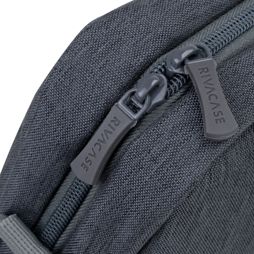 Primo piano di due cerniere grigie su una borsa in tessuto grigio scuro. Entrambe le cerniere riportano la scritta 'RIVACASE'.