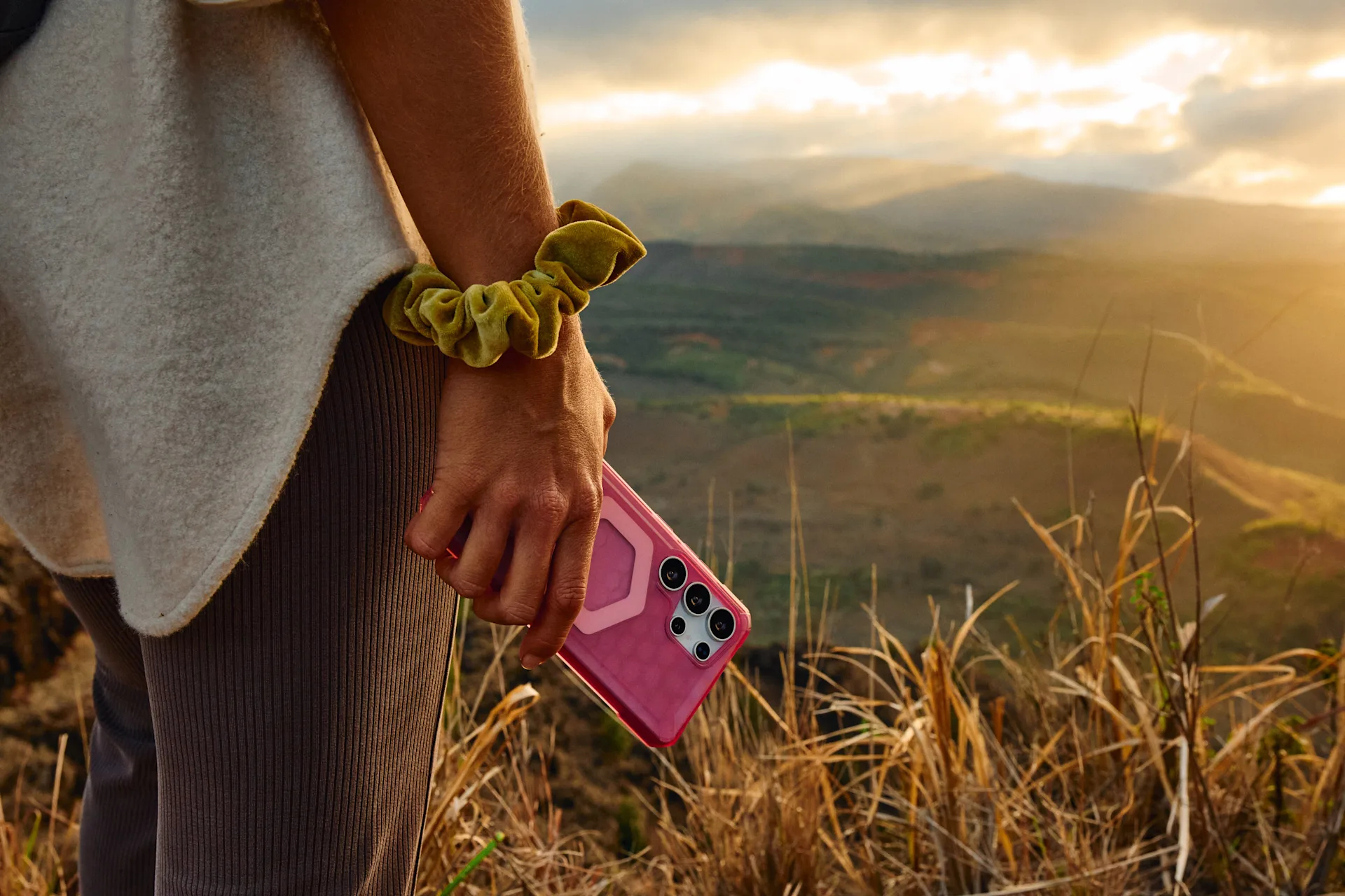 Eine Hand hält ein pinkes Handy vor einer bergigen Landschaft bei Sonnenuntergang.