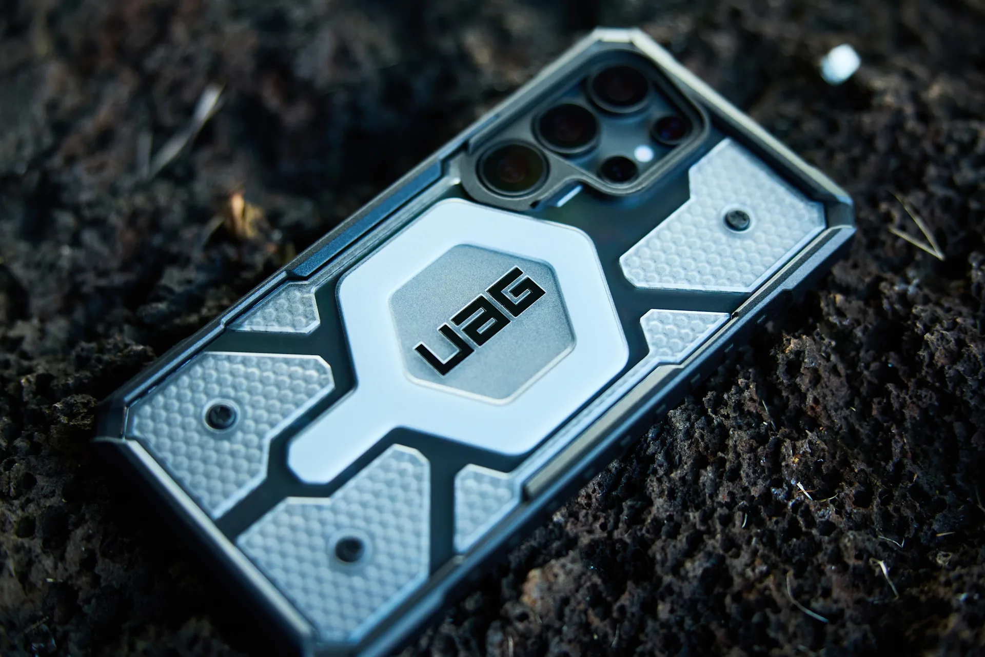 Una custodia per telefono UAG nera e argento con logo esagonale poggia su un terreno scuro e texturizzato.