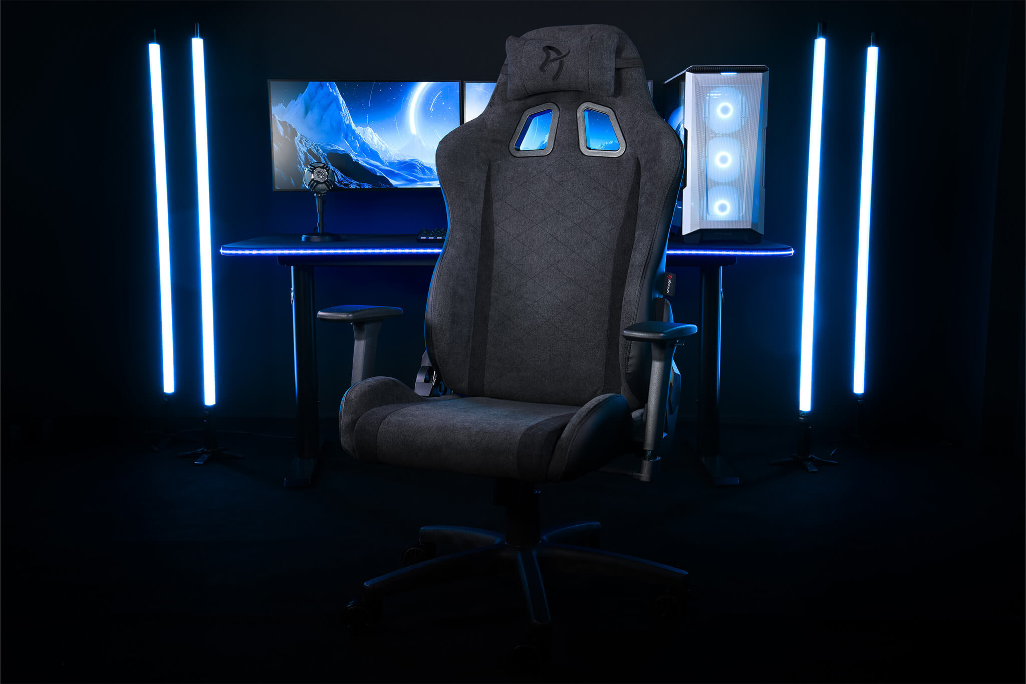Una sedia da gaming scura davanti a una scrivania con monitor e PC. Luci blu.