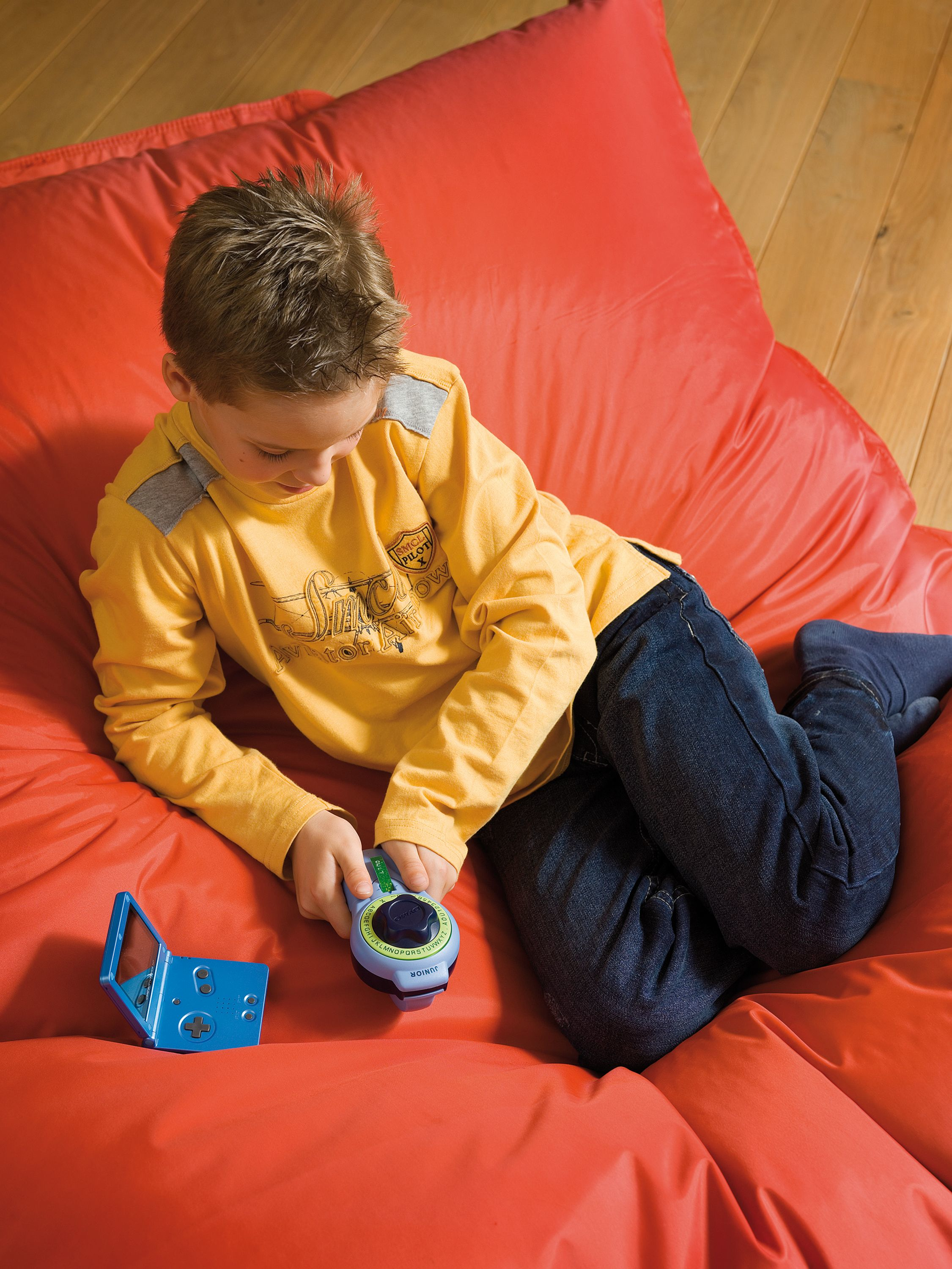 Junge mit gelbem Pullover und blauen Jeans sitzt auf einem roten Sitzsack und spielt.