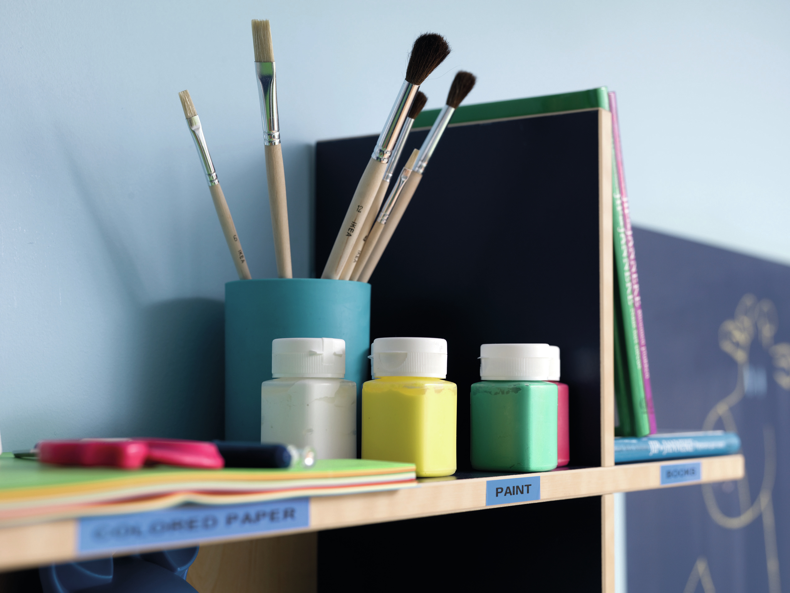 Pinceles en un portaobjetos azul junto a botes de pintura, libros y papel.