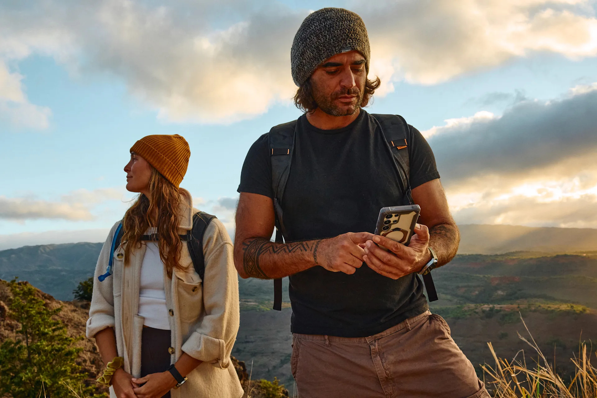 Hombre con móvil y mujer con gorro en una colina al atardecer.