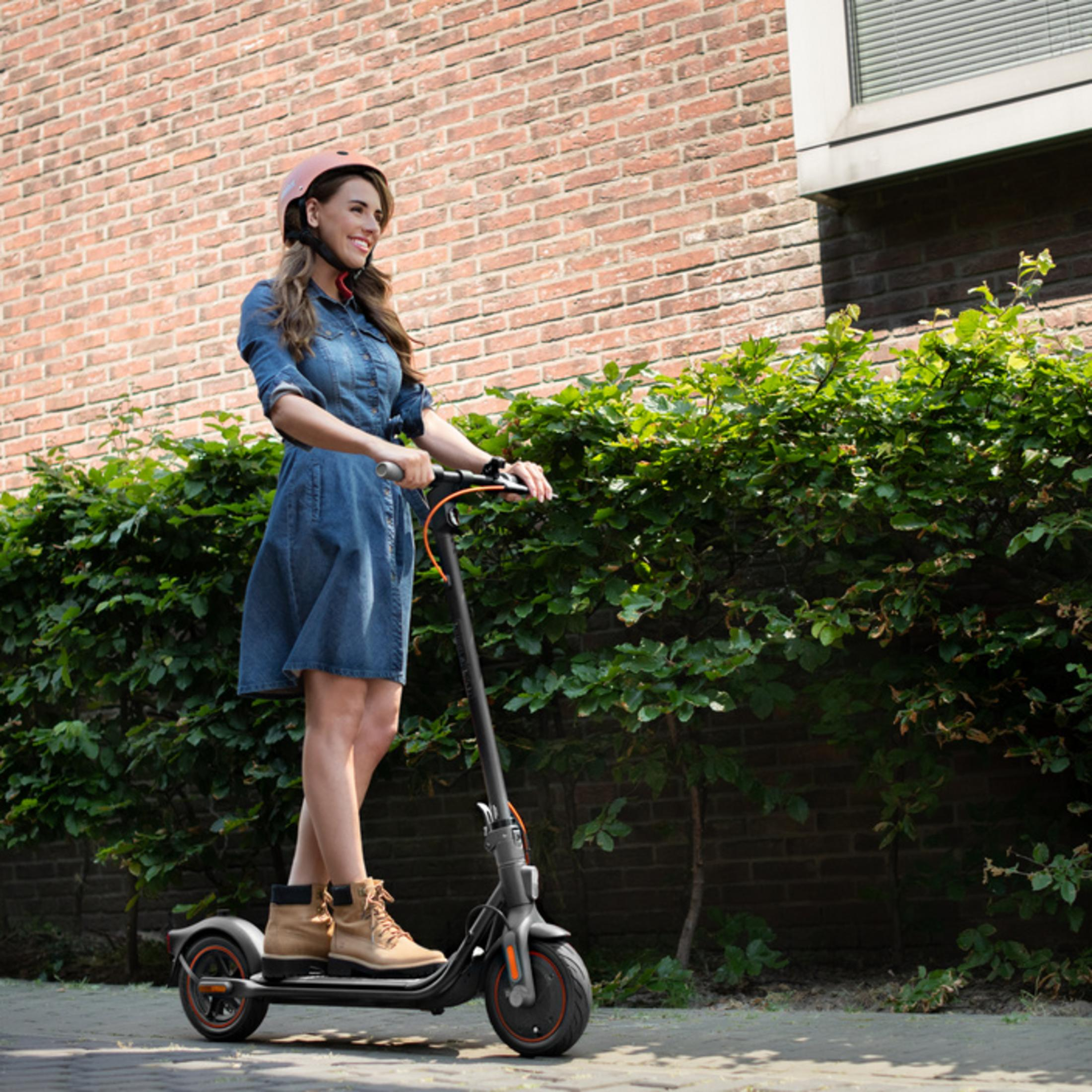 Eine Frau in einem Jeanskleid fährt einen E-Scooter, trägt einen Helm und steht vor einer Ziegelwand.