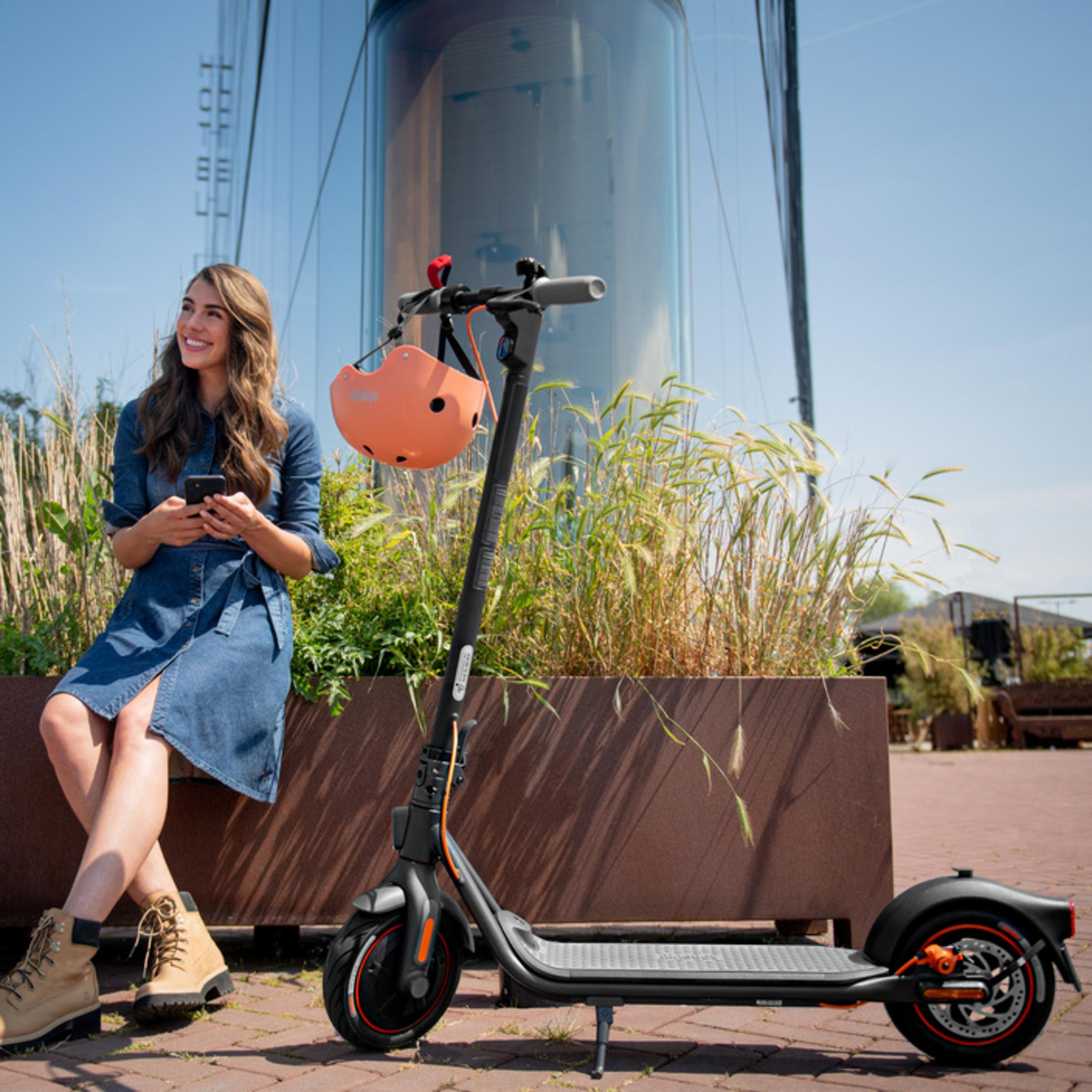 Eine Frau sitzt auf einer Bank, neben ihr ein E-Scooter mit Helm. Draußen an einem Gebäude.