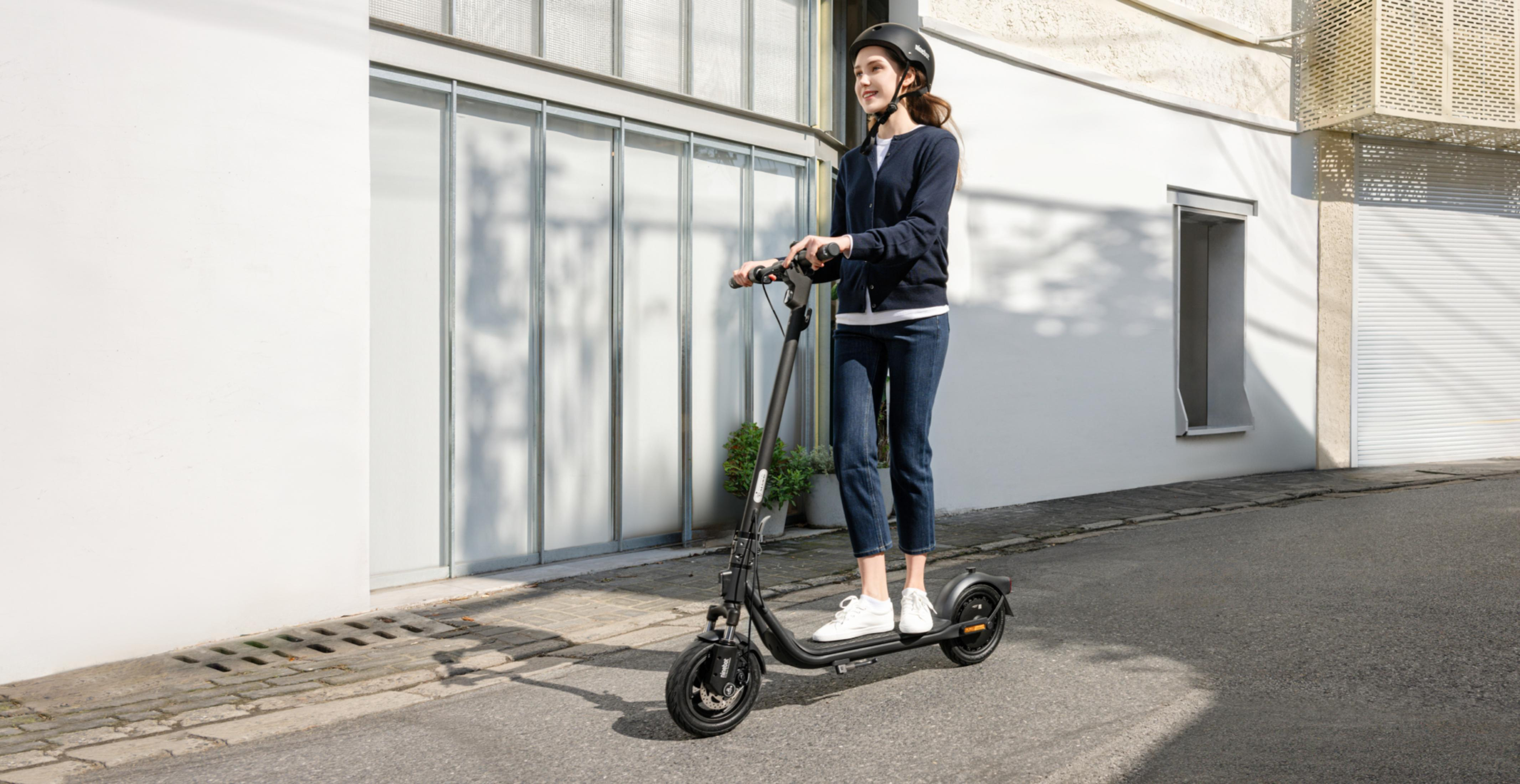 Eine Frau mit Helm fährt einen schwarzen Elektroroller auf einer Straße.
