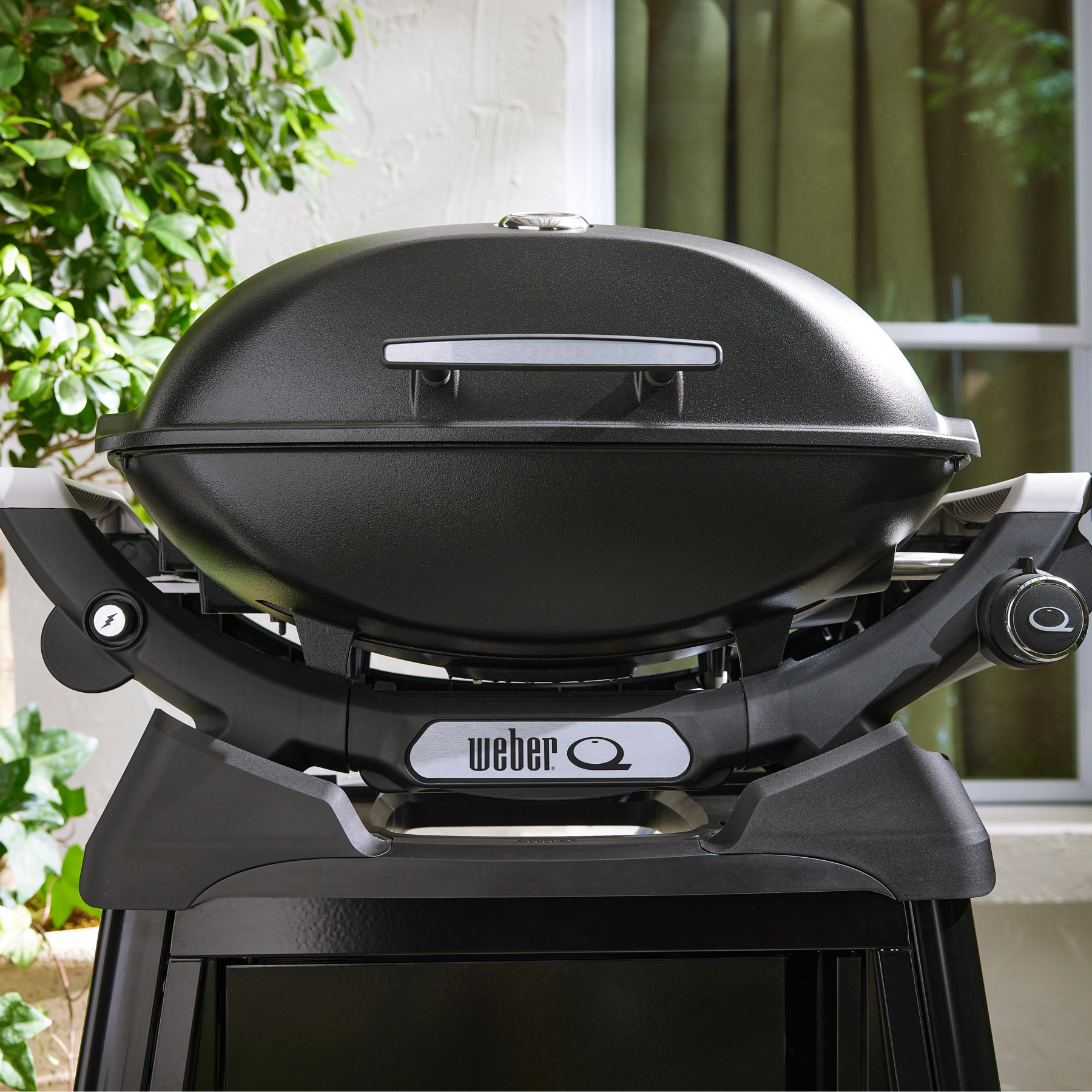 Una griglia Weber nera con manico argentato davanti a una finestra e piante.