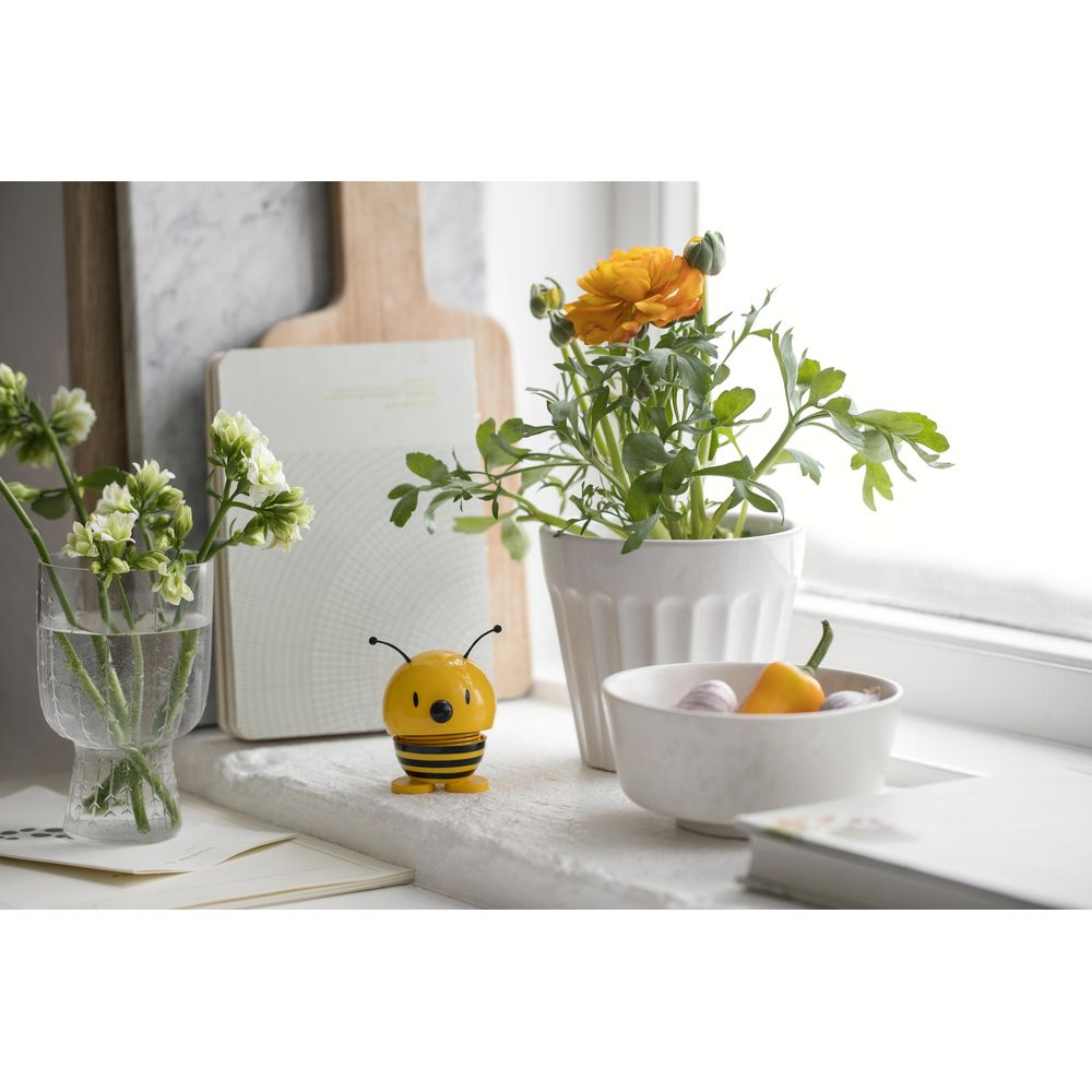 Eine Biene-Figur steht neben einer Pflanze mit einer orangefarbenen Blüte und einer Schale mit Knoblauch und Peperoni.
