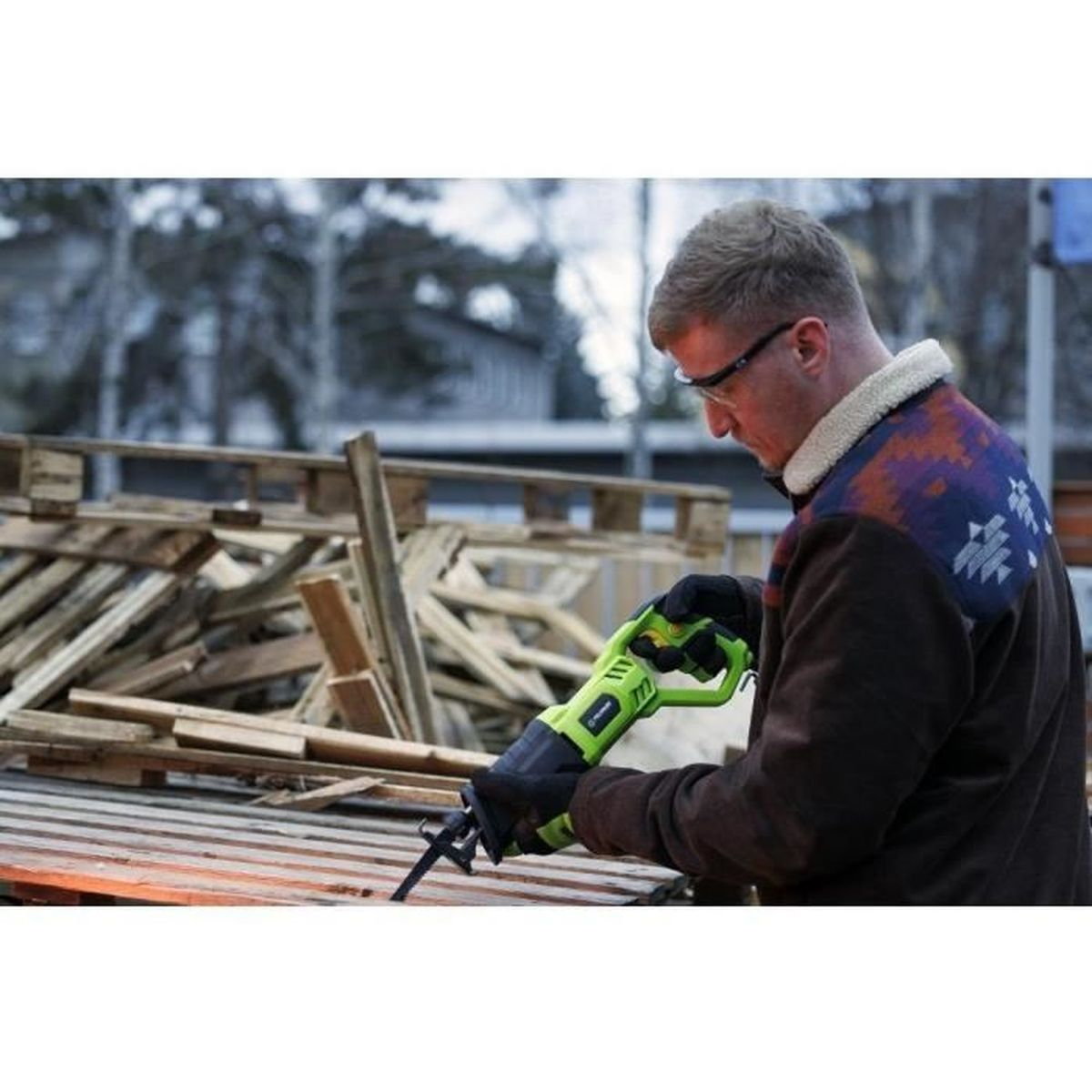 Mężczyzna w okularach ochronnych piłuje drewno piłą szablastą. Drewniane palety w tle.