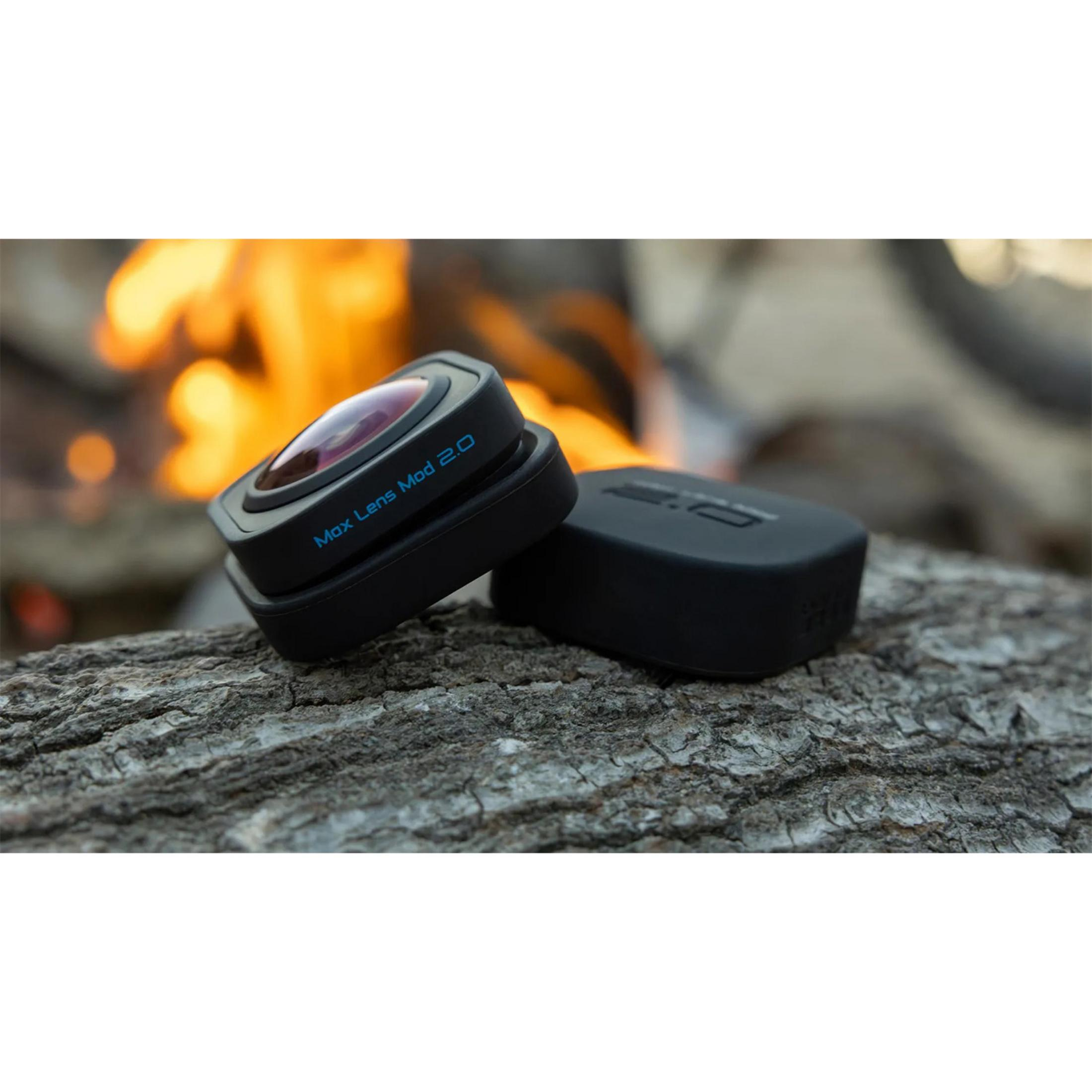 Ein schwarzer Objektivaufsatz mit der Aufschrift „Max Lens Mod 2.0“ liegt auf Holz neben einem Feuer.