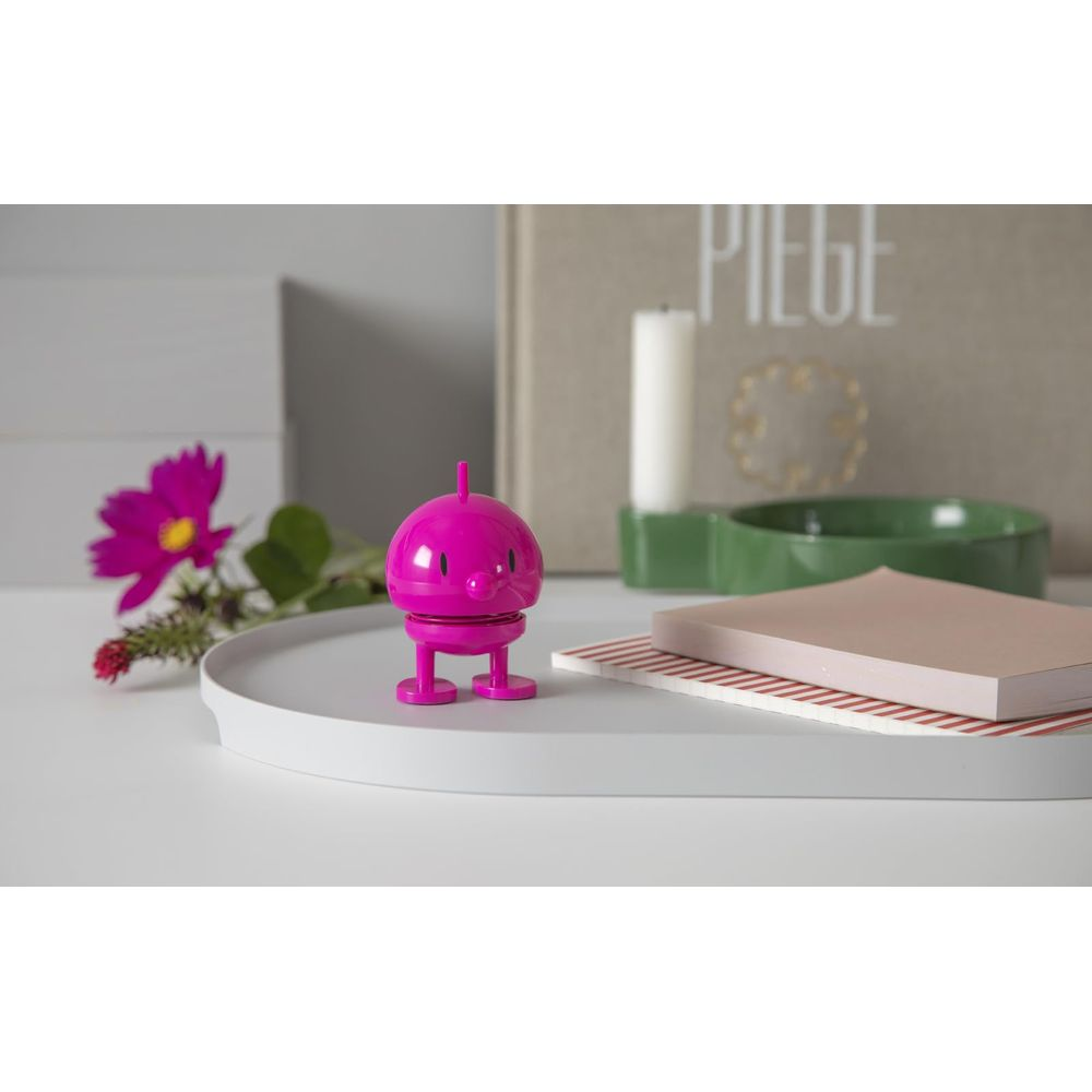 Ein kleiner pinker Roboter steht auf einem Tablett neben einer Blume und Büchern.
