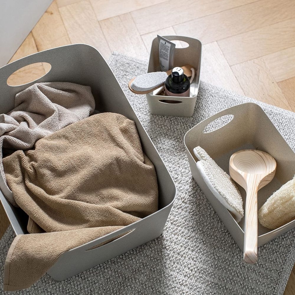 Drei beige Aufbewahrungsboxen auf einem Teppich. Eine enthält Handtücher, eine Badausstattung, eine Toilettenartikel.