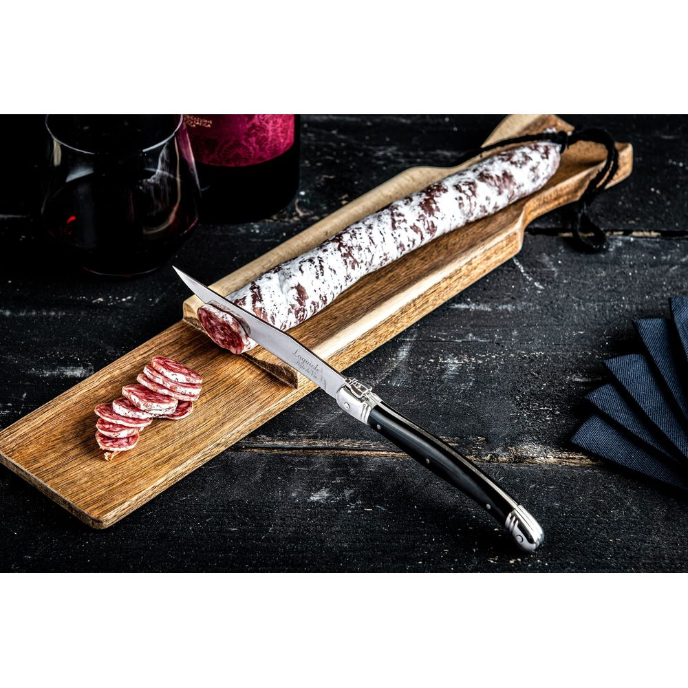 Ein Laguiole-Messer schneidet eine Trockenwurst auf einem Holzbrett mit Wein und dunklen Servietten.