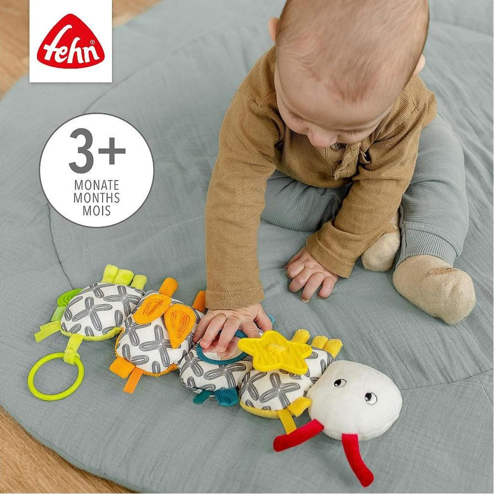 Baby spielt mit buntem Greifspielzeug auf einer grauen Matte. Alter 3+.