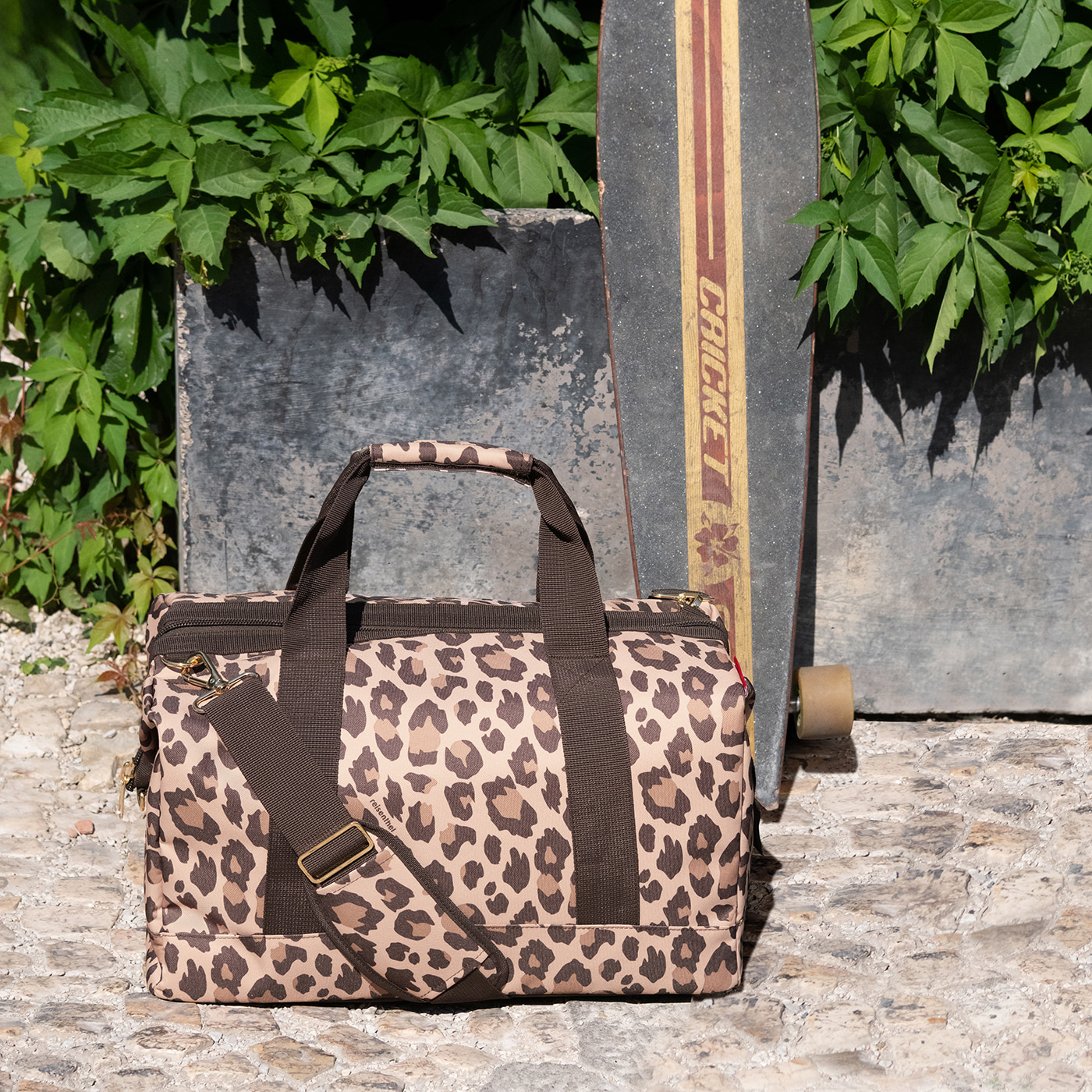 Eine leopardgemusterte Tasche steht neben einem Skateboard vor grünem Laub.