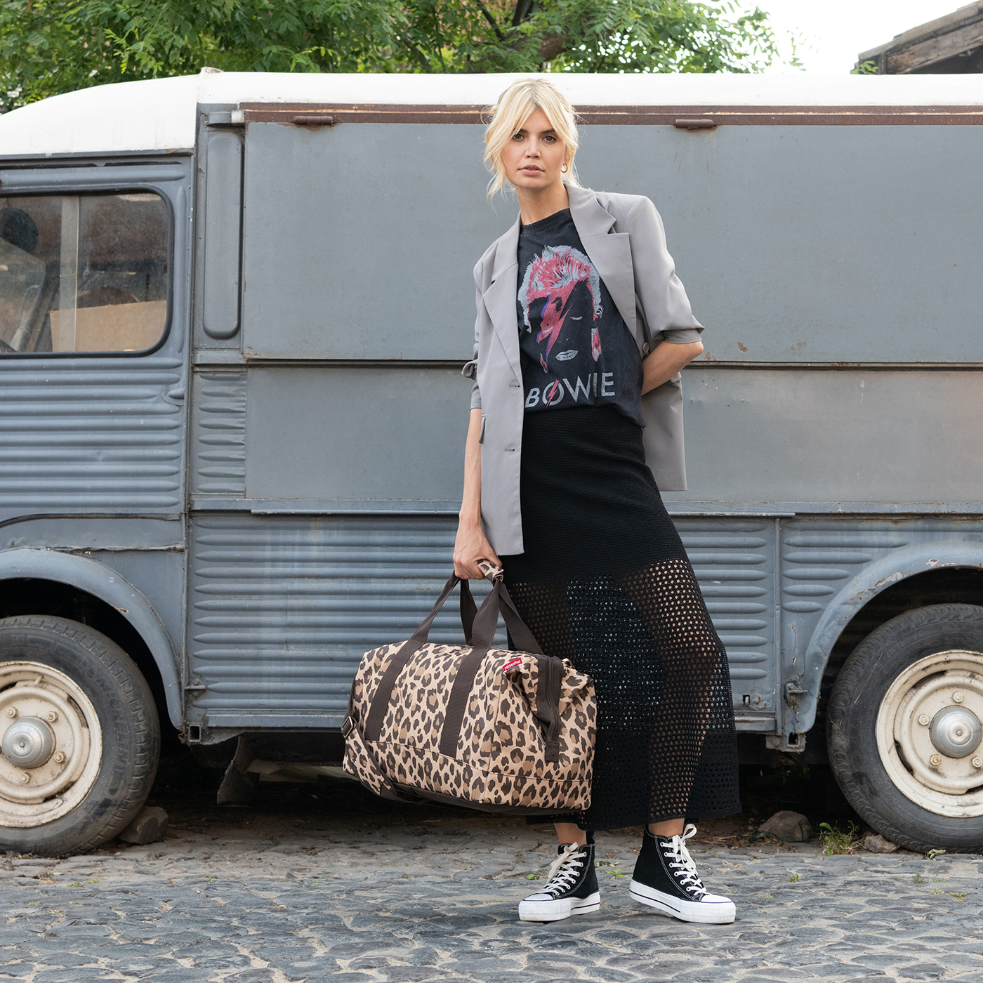 Eine Frau mit blonden Haaren steht vor einem grauen Lieferwagen und hält eine Leoparden-Tasche.