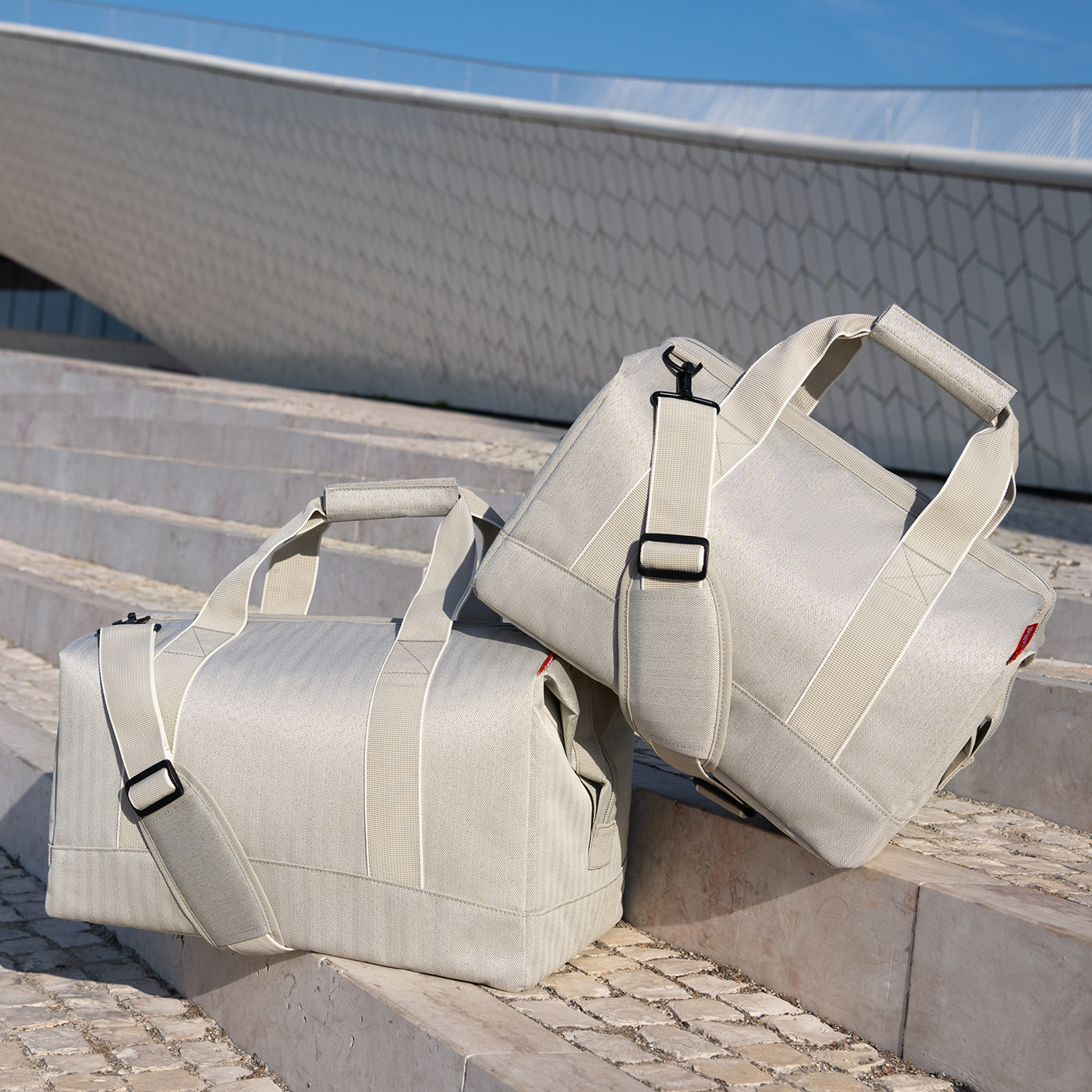Zwei hellgraue Reisetaschen stehen auf einer Steintreppe vor einem modernen Gebäude.