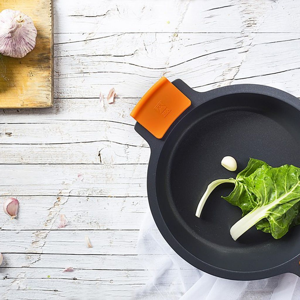 Graue Pfanne mit orangem Griff, Knoblauch und grünem Blattgemüse auf Holztisch.