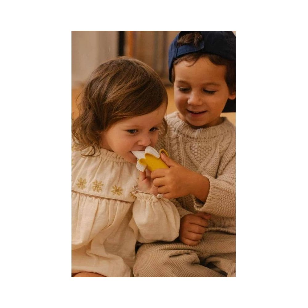 Ein Junge bietet einem Mädchen einen gelben Bananenbeißring an. Beide sitzen.