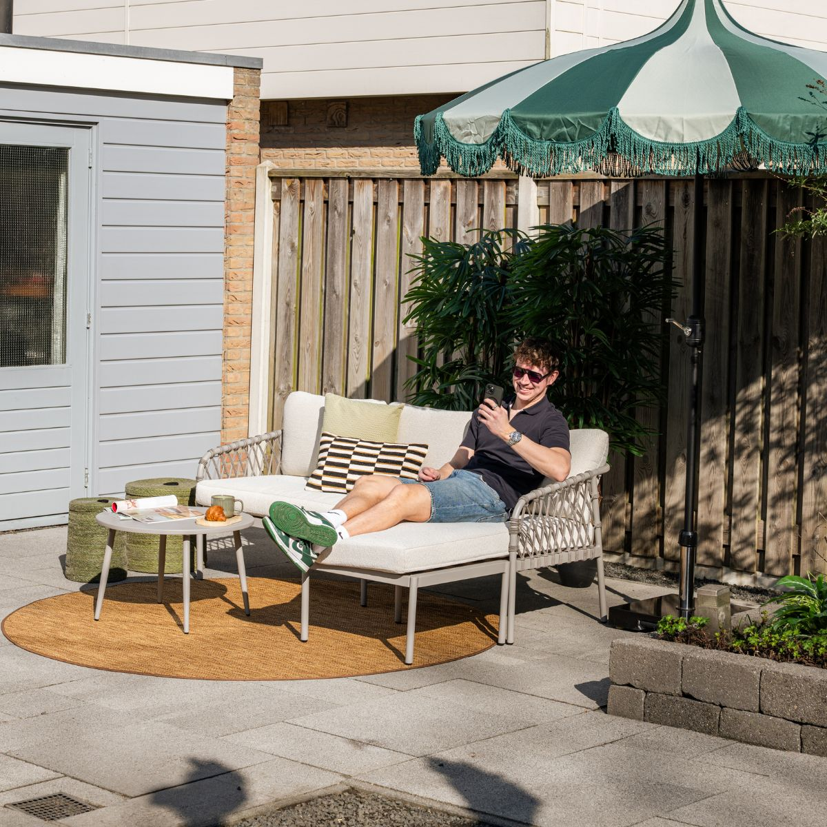 Man on outdoor sofa using phone, with umbrella and table nearby.