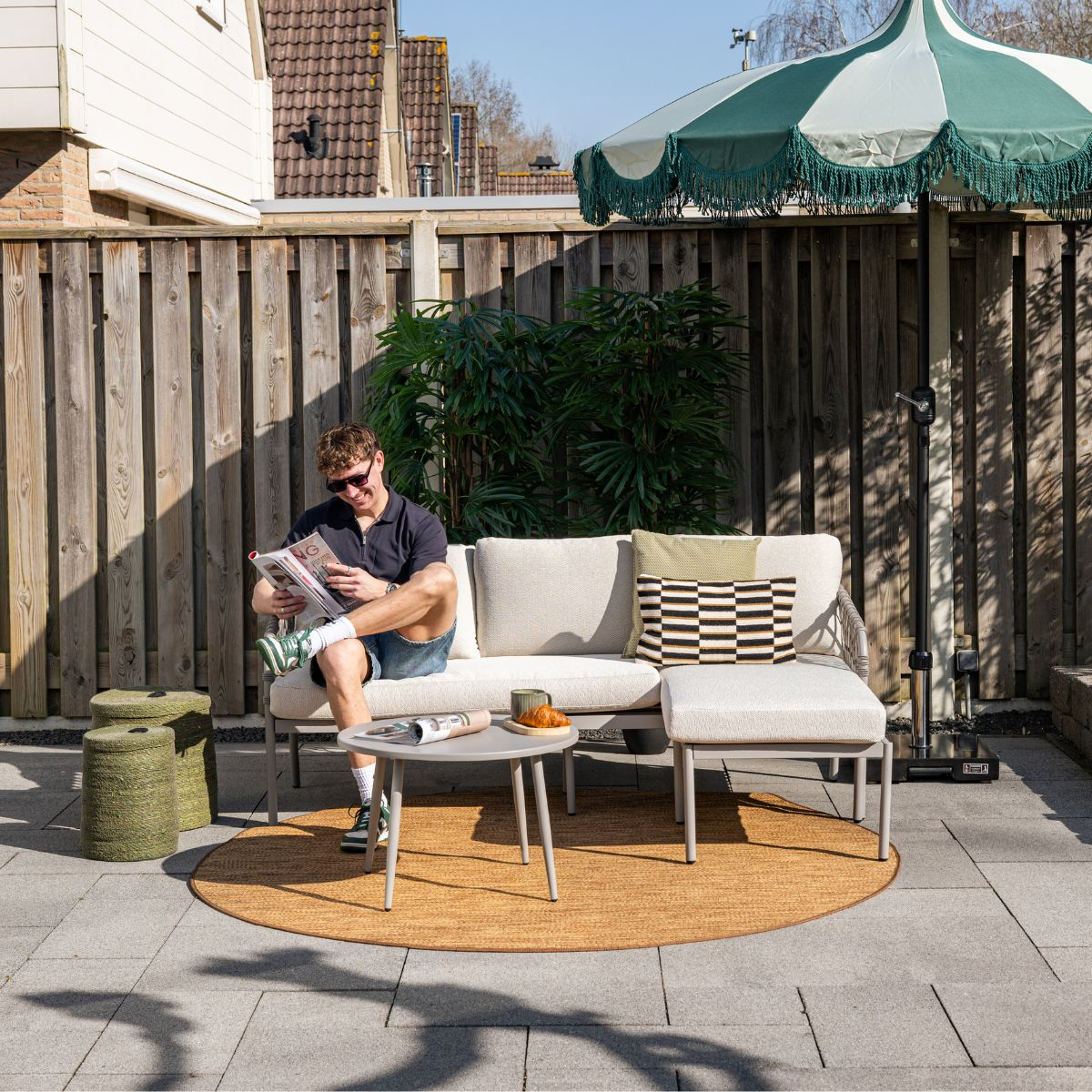 Man reading magazine on outdoor sofa, green stools, coffee table, umbrella.