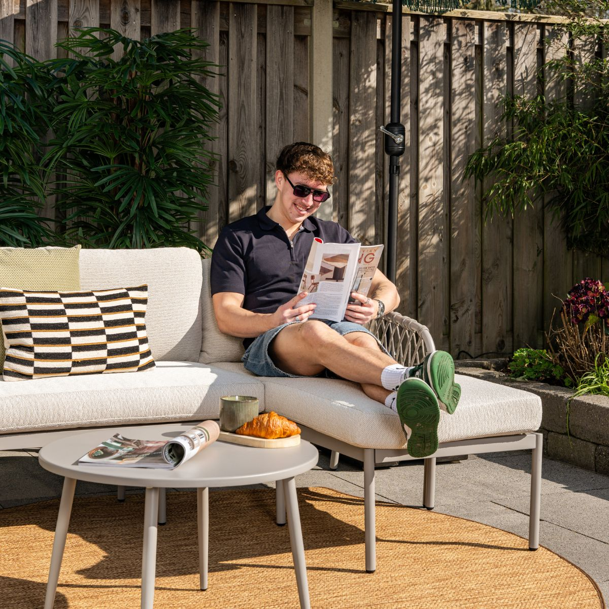 A man reads a magazine on an outdoor sofa. A croissant is on the table.