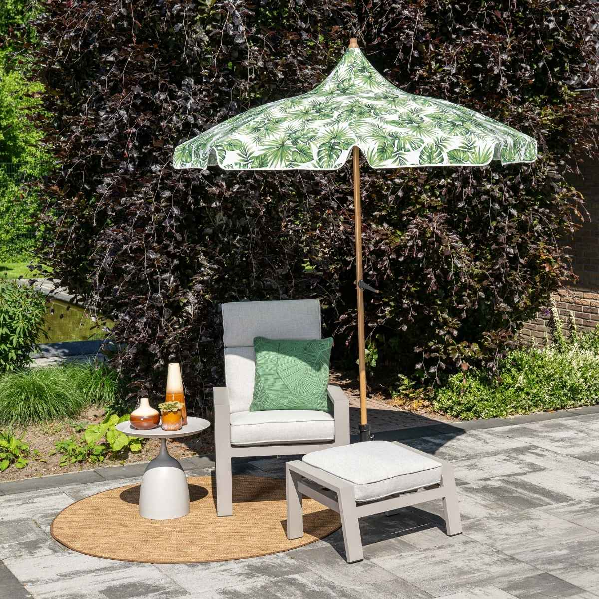 Een stoel met ottoman staat onder een paraplu met tropisch bladerpatroon op een patio.