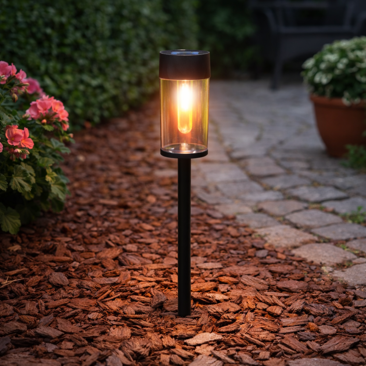 Garden light with flame effect next to blooming flowers and a stone path.