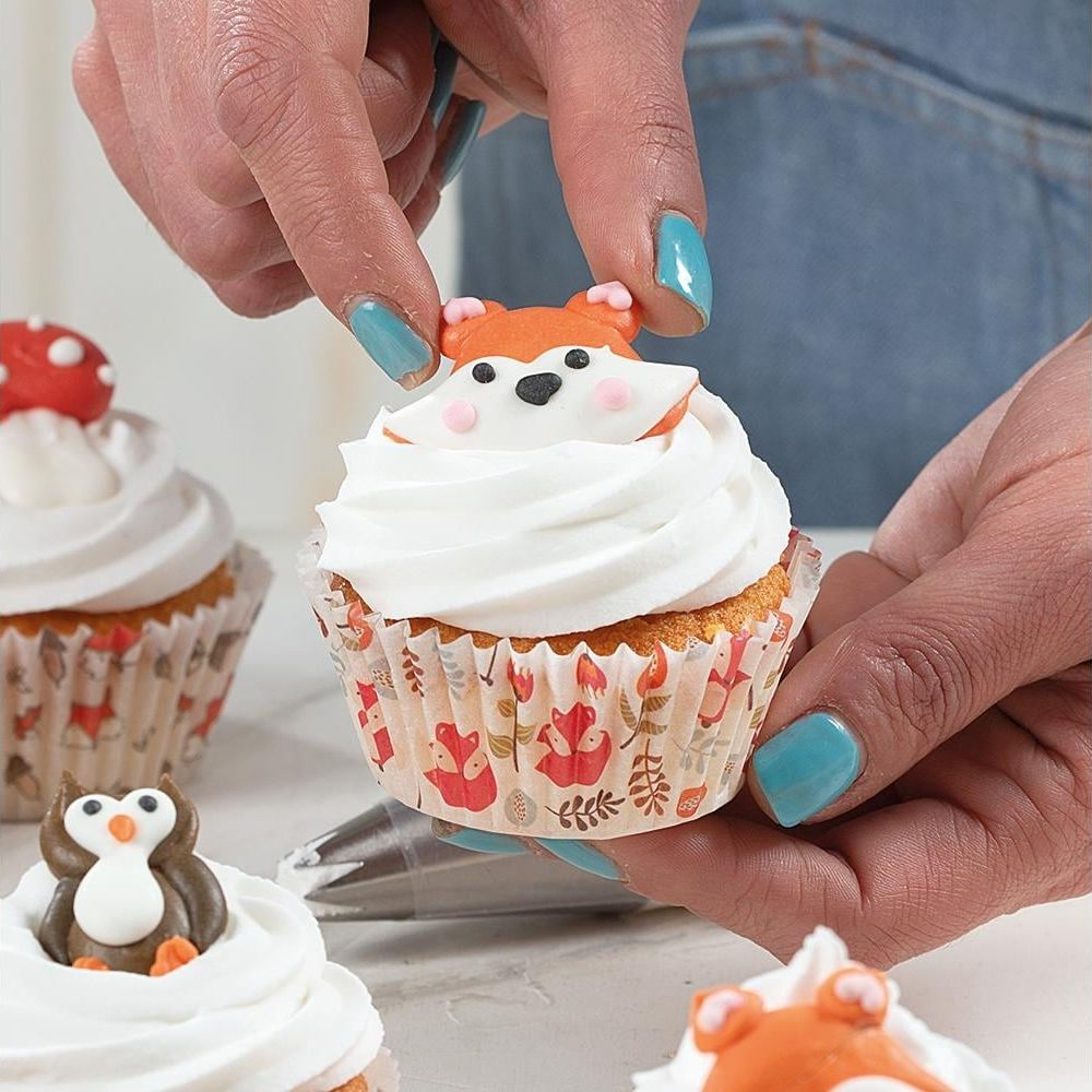 Eine Hand platziert eine Fuchs-Dekoration auf einem Cupcake mit weißer Glasur.