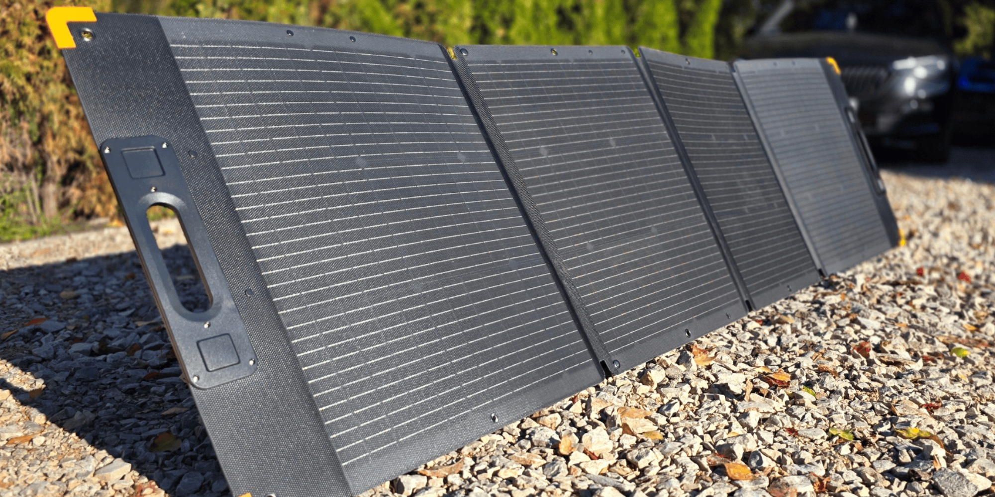 Składany panel słoneczny leży na żwirze, z samochodami i drzewami w tle.