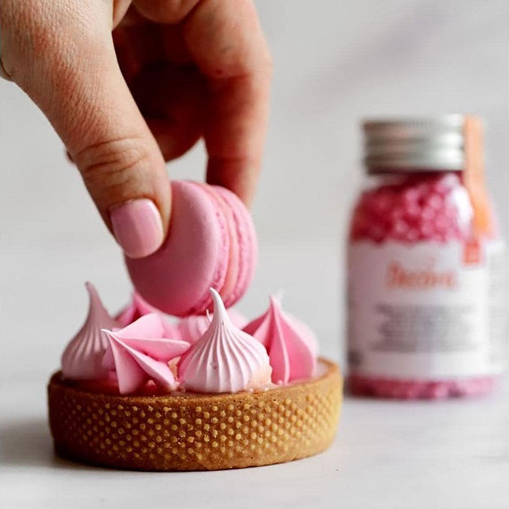 Eine Hand platziert eine rosa Macaron auf einem Tartelette mit rosa Baiser.