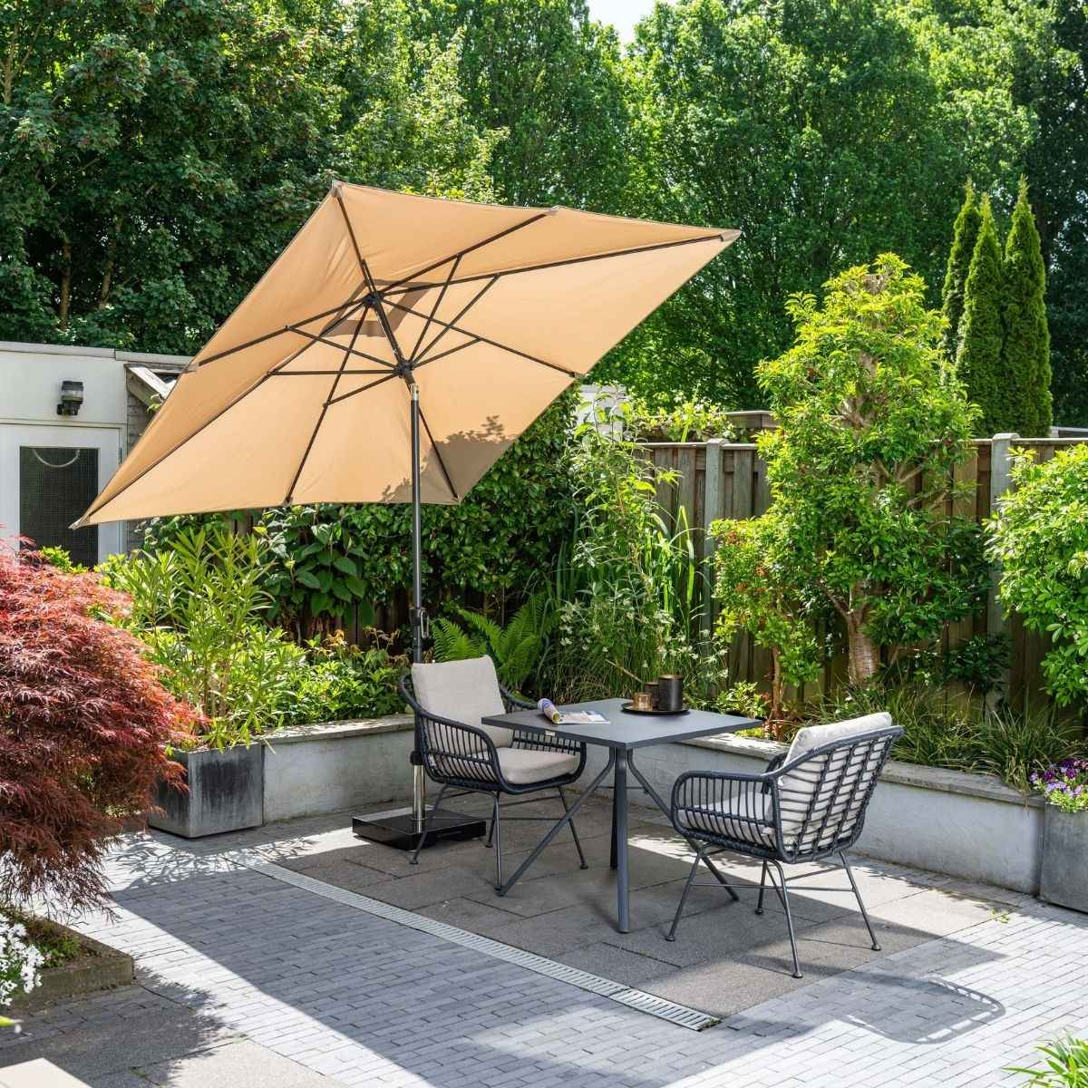 Een terrasmeubelset met beige parasol, tafel en stoelen, omringd door weelderige groen.