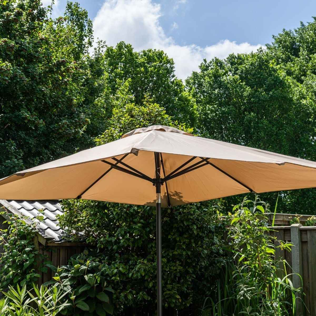 Een grote, beige parasol staat open in een tuin met bomen en een hek.