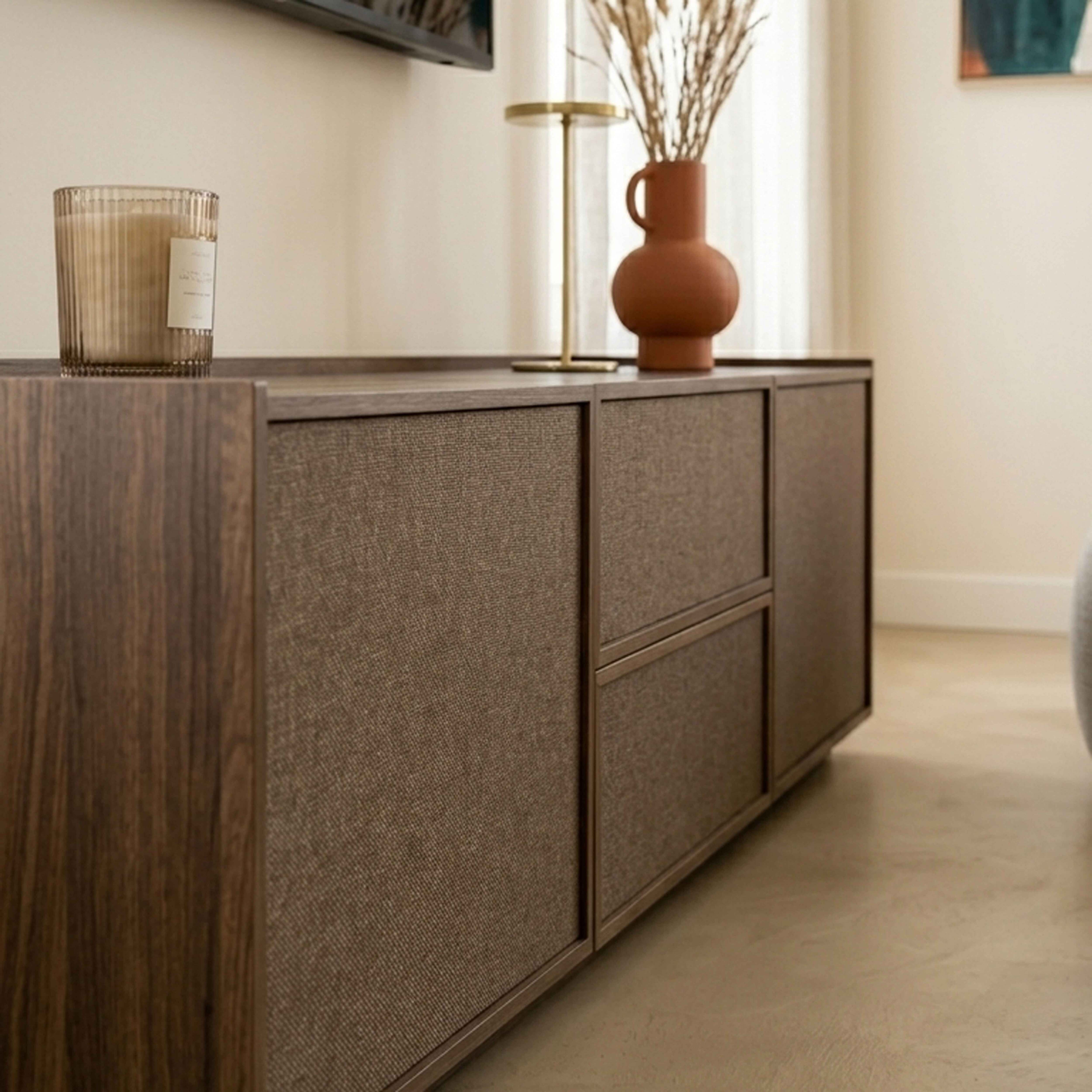 Dunkelbraunes Sideboard mit Stofffronten, Kerzenhalter und Vase mit Trockenblumen.