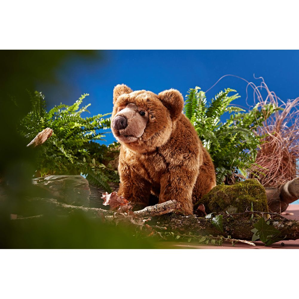 Ein brauner Teddybär sitzt auf einem moosbewachsenen Ast vor Farnen und blauem Himmel.