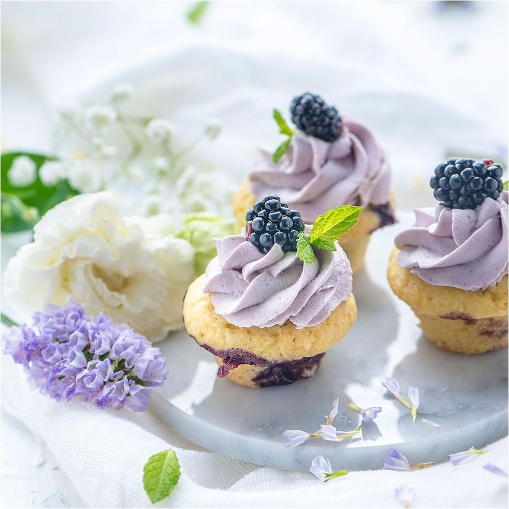 Drei Cupcakes mit lila Zuckerguss, Brombeeren und Minze auf Marmorplatte.