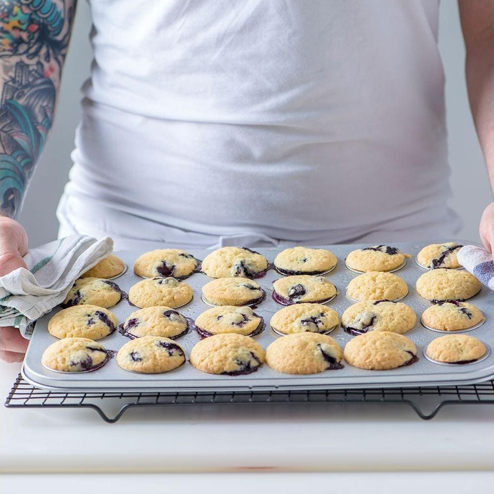 Ein Mann mit einem T-Shirt hält eine Blechform mit 20 Blaubeer-Muffins auf einem Gitter.