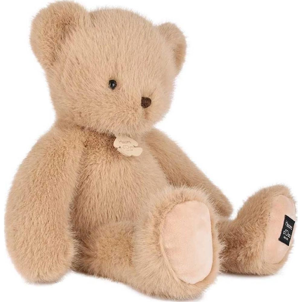 Un orsacchiotto di peluche marrone chiaro e soffice è seduto con le gambe distese. Ha una piccola etichetta sul petto.