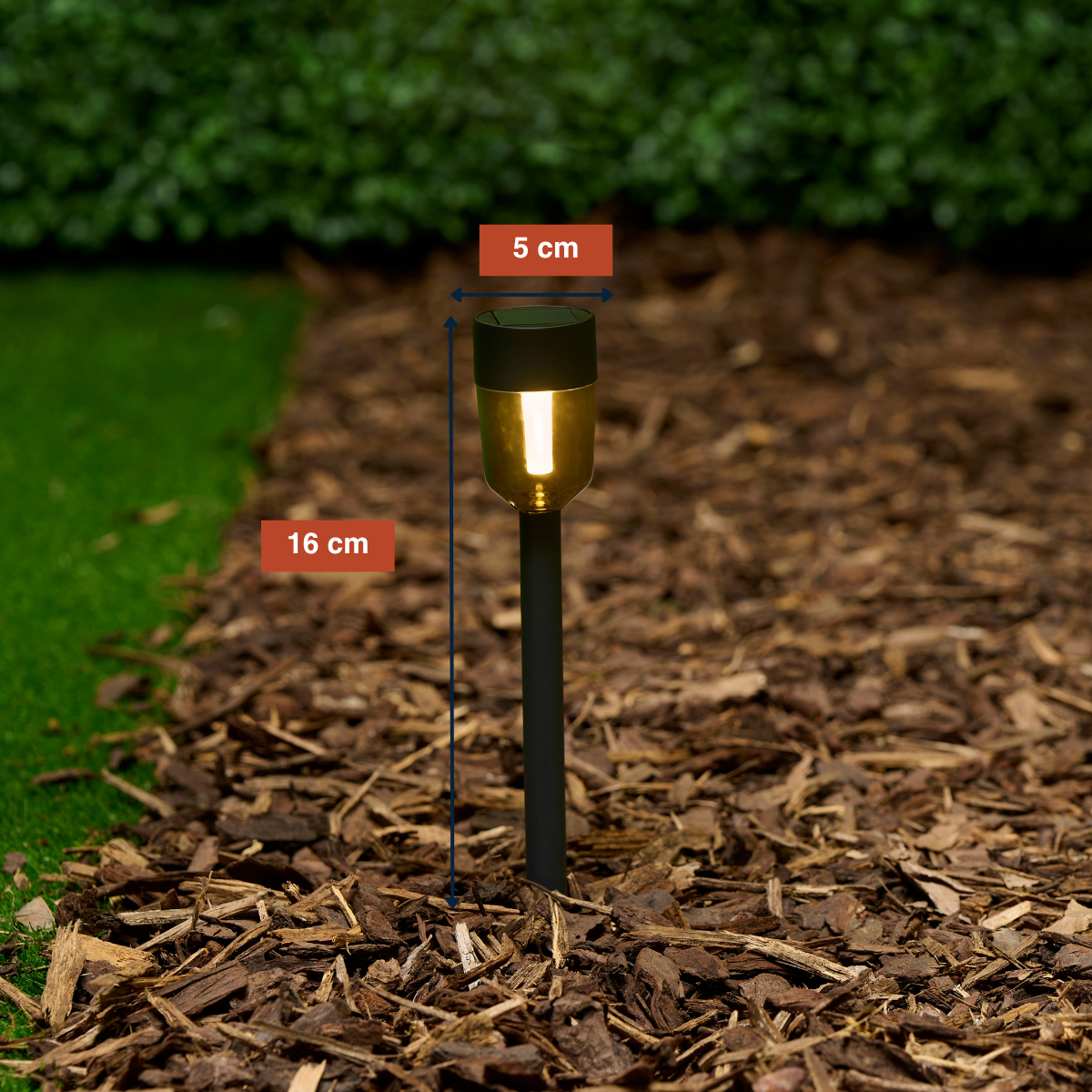 A 16cm tall solar garden light with a 5cm width, lit up in bark mulch.
