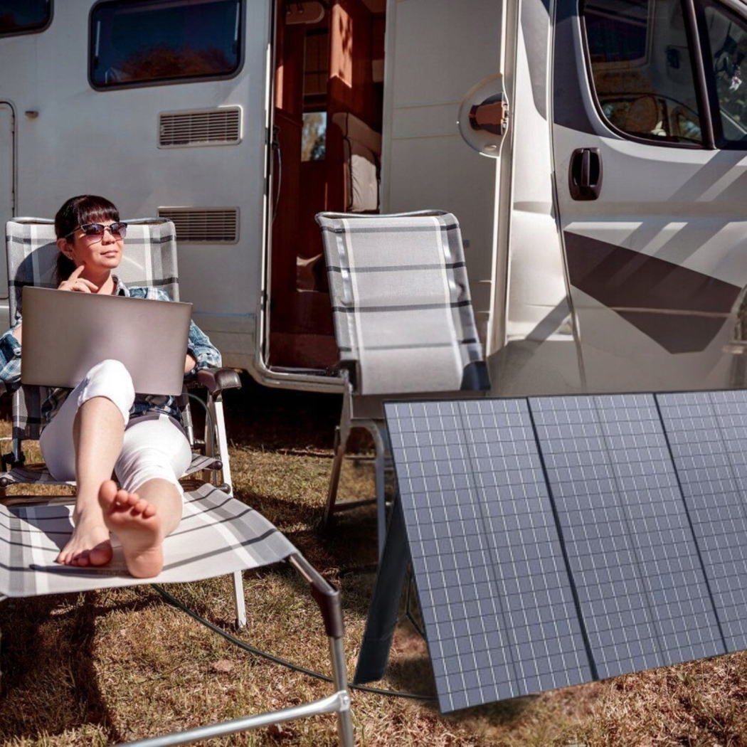 Kobieta siedzi w krześle na zewnątrz, używając laptopa obok kampera.